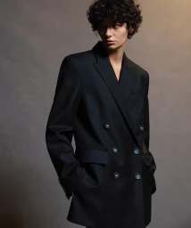 Model with curly dark hair wearing a structured black double-breasted blazer against a textured grey studio background.