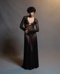 Model in a long black sheer lace dress standing against a textured grey studio background for Common Hours.