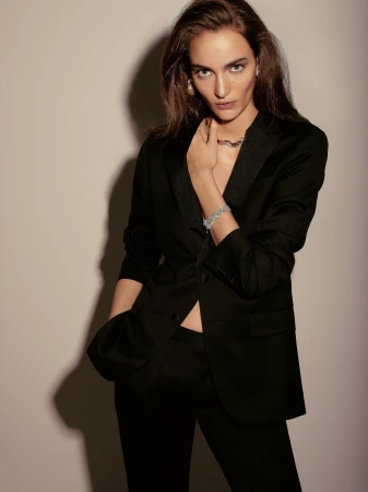 Woman in a tailored black suit and diamond jewelry posing against a neutral studio backdrop with directional shadow.
