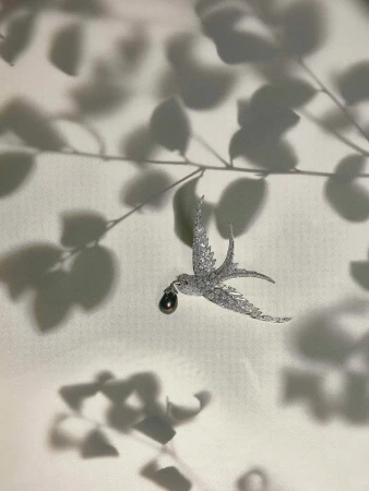 Sparkling bird brooch with a dark pearl against a pale background with leaf shadows, creating an elegant and visually intriguing piece of jewellery.
