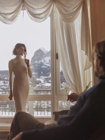 Woman in white dress holds champagne glass by a window overlooking snow-capped mountains while a man sits in the foreground.