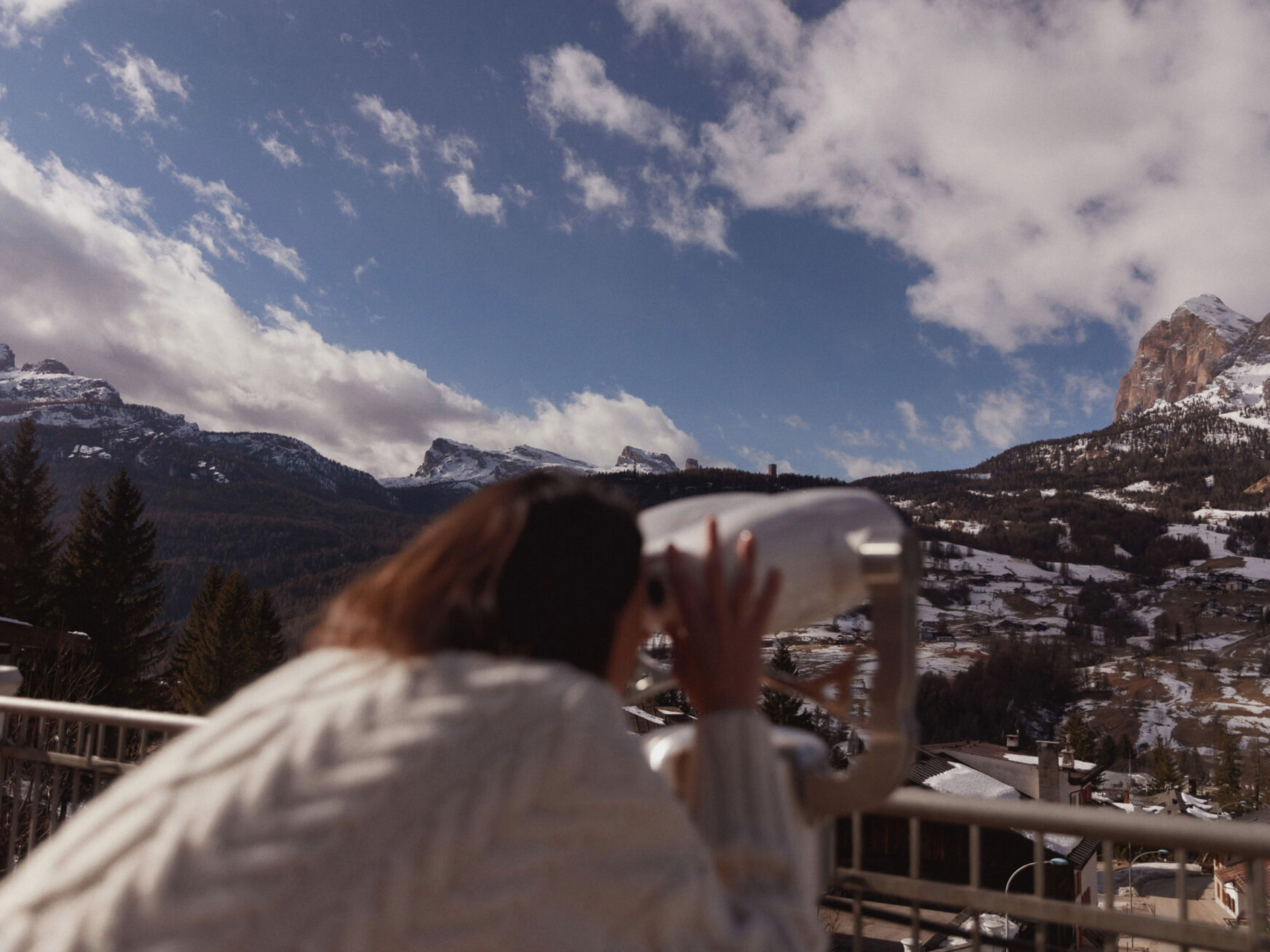 Adult female in a white sweater using binoculars overlooking a snowy mountain range with a blue cloudy sky backdrop during daytime.