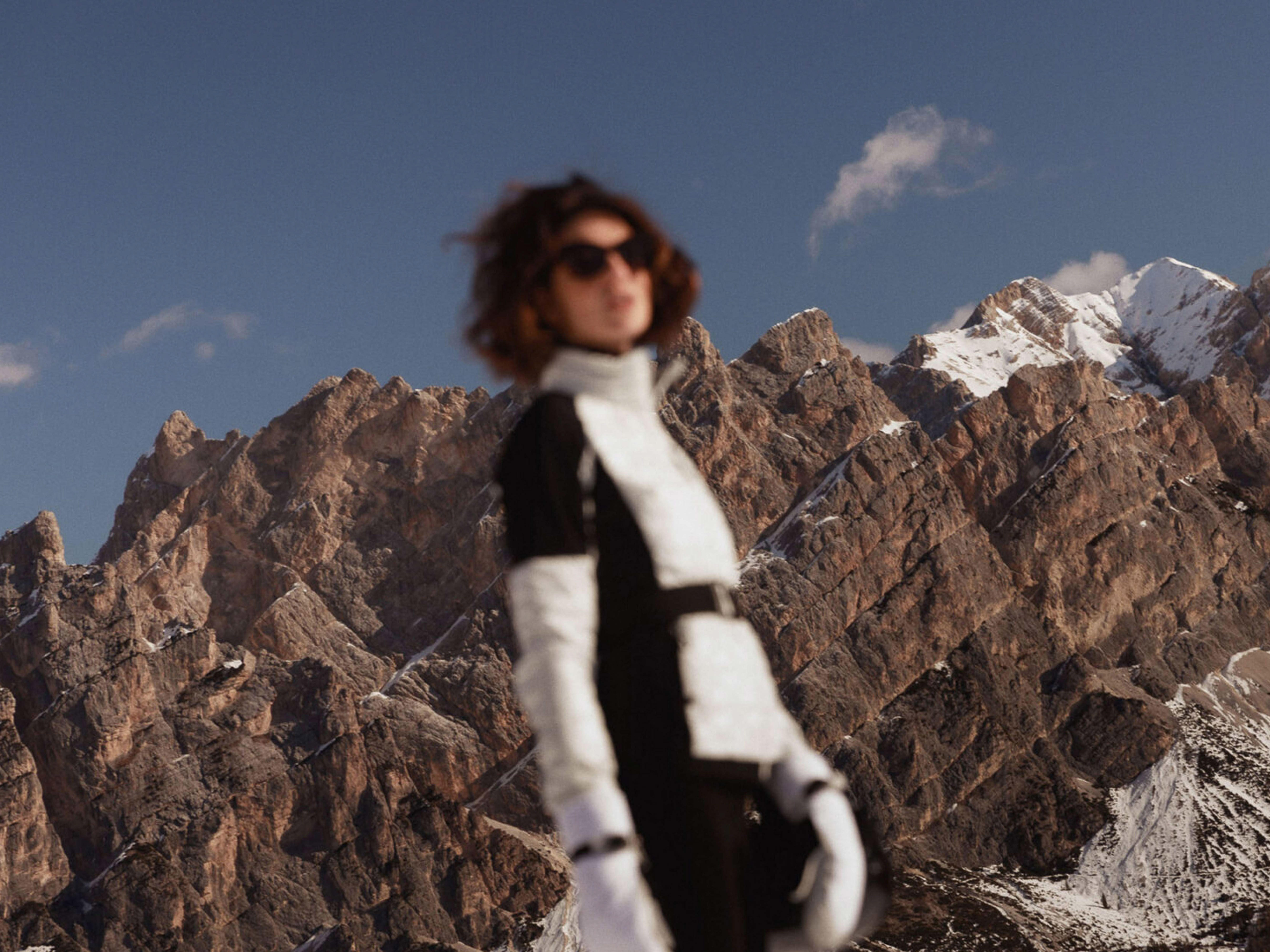 Woman in ski suit against mountain backdrop with snow-capped peaks under blue sky. Outdoor fashion shot on sunny day.