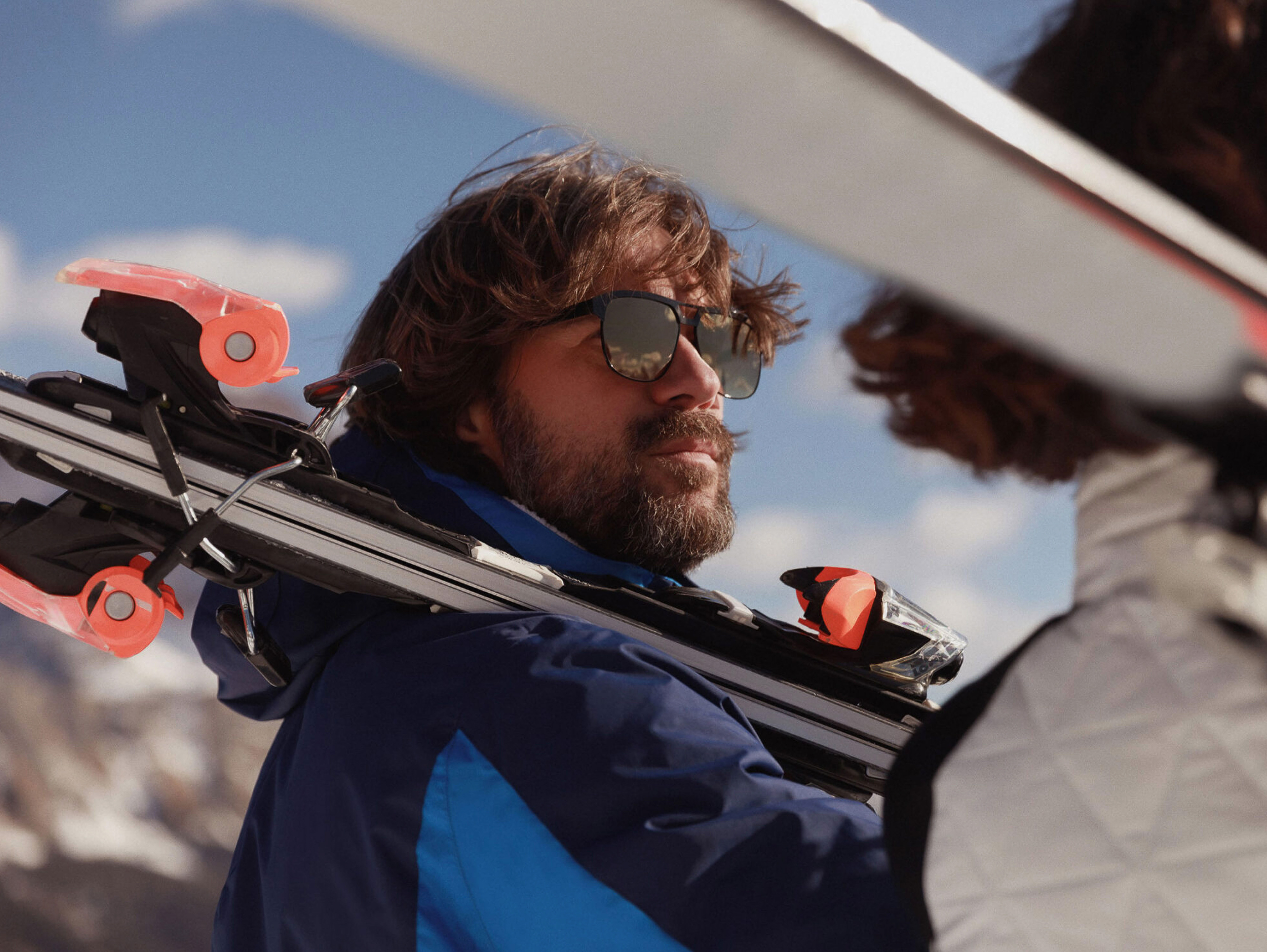A man with sunglasses and beard carries skis over his shoulder, standing near a woman on a sunny day in a mountainous, snowy landscape.