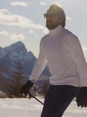 Man in white turtleneck sweater skis on snowy mountain with sunglasses, gloves, and poles under bright sky with mountains in background.