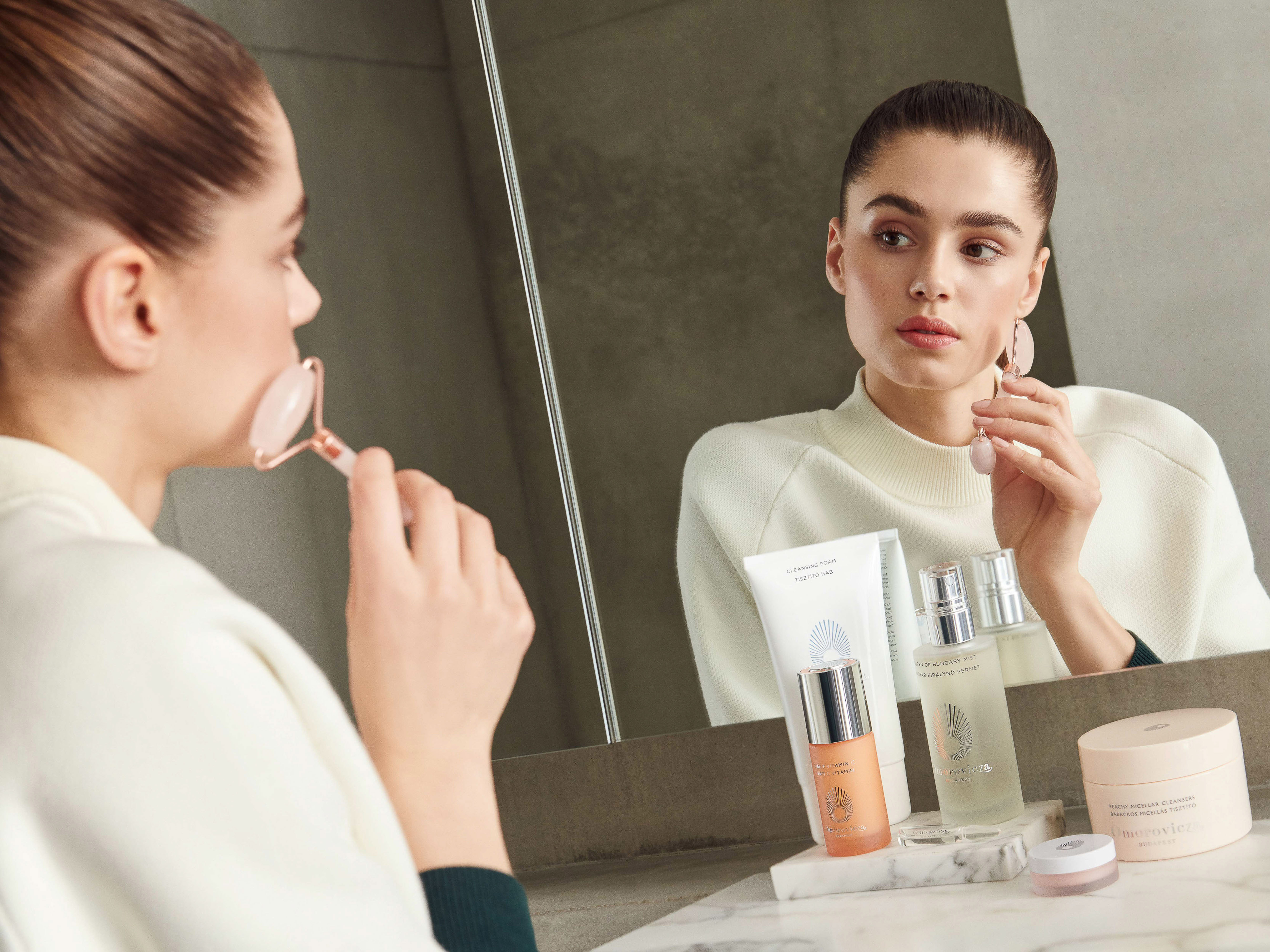 Adult woman using a rose quartz face roller in front of a mirror with skincare products in a beauty and wellness setting.