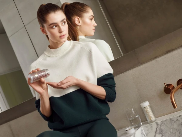 Indoor beauty shot features a young woman in sportswear, holding a pill bottle, with a reflective background and natural light.