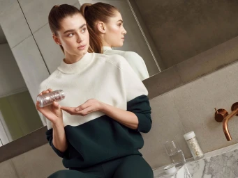 Woman in a green and cream sweater holding a bottle of supplement capsules in a modern bathroom with a mirror reflection.