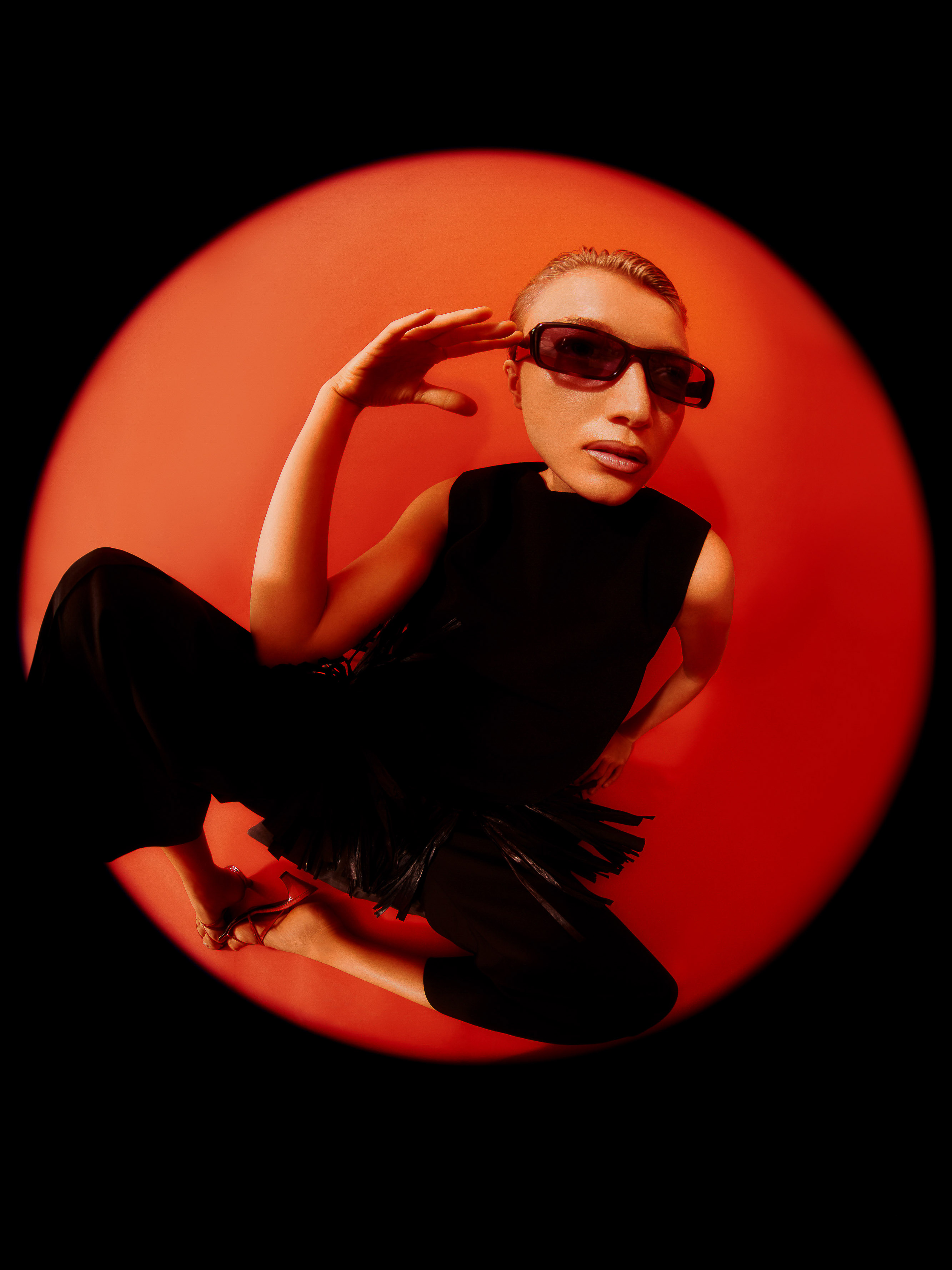 Woman in sunglasses poses in black against a red background with a wide angle lens, creating a fisheye effect and a circular vignette.