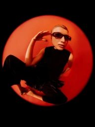 High-angle shot of a model wearing dark rectangular sunglasses and a black sleeveless outfit framed within a red circular spotlight.
