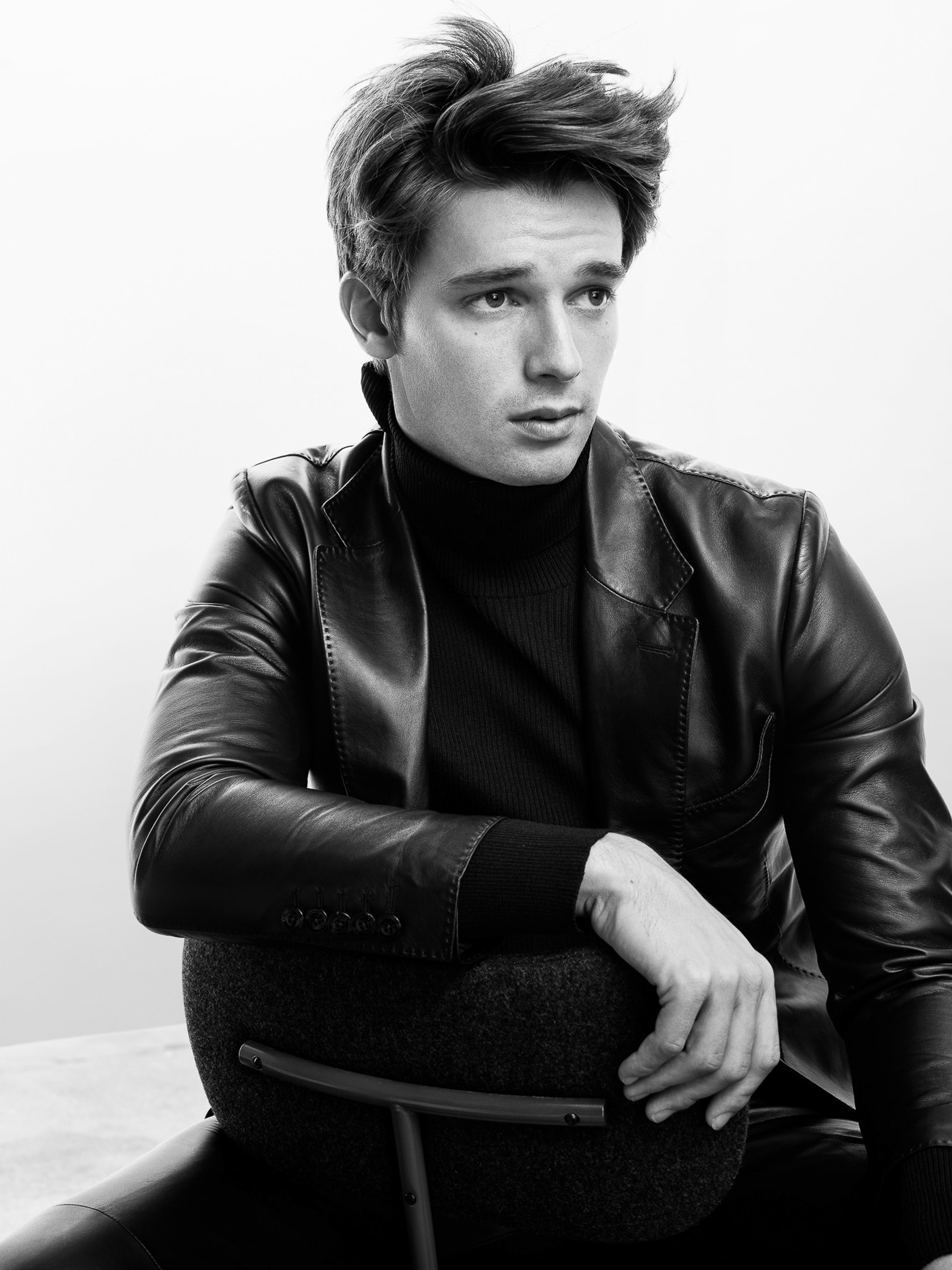 Black-and-white portrait of a young adult man wearing a leather jacket and turtleneck, sitting on a chair against a white background.