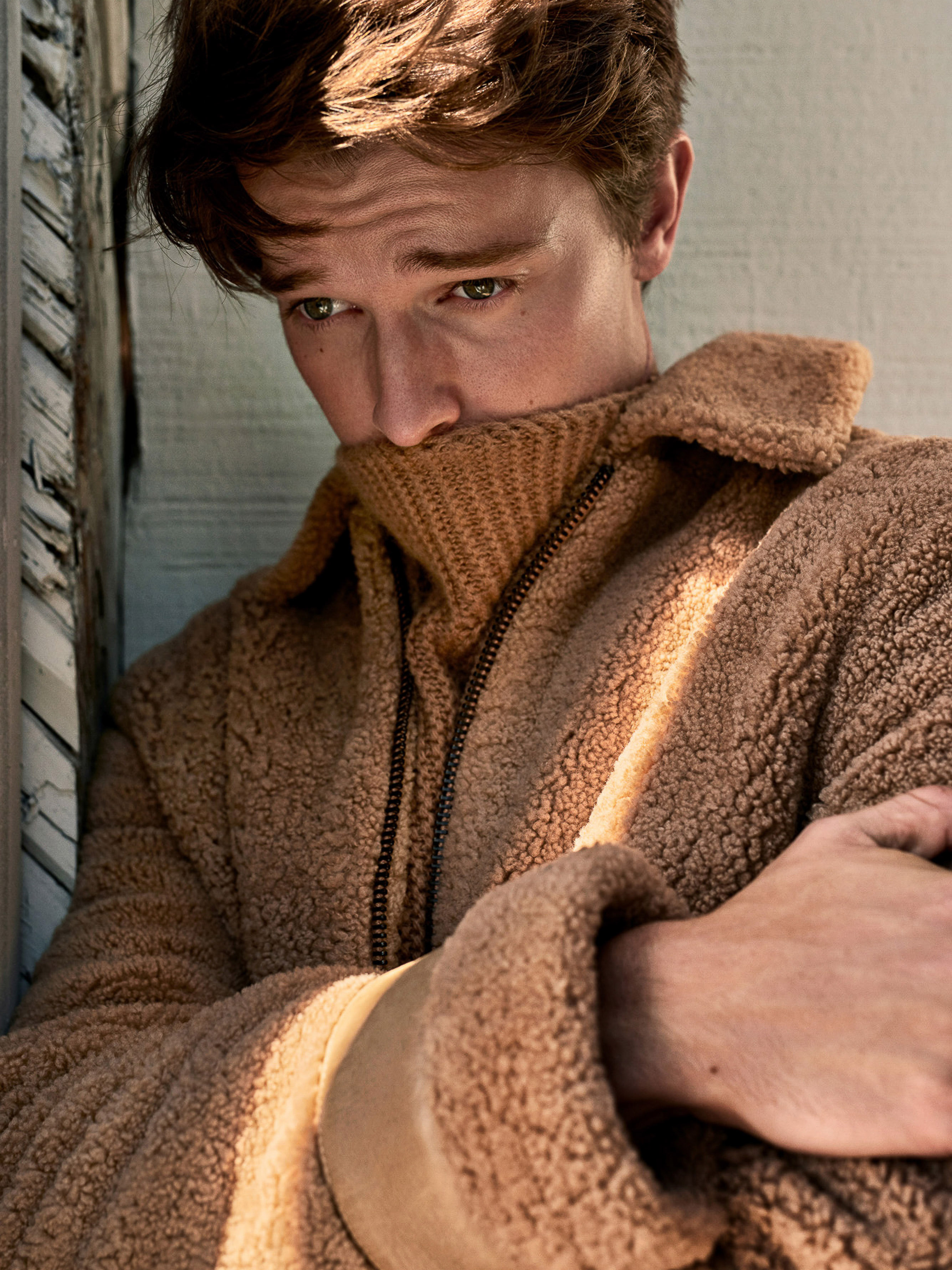 A young adult male in a light brown teddy coat pulls up a tan turtleneck sweater, posing in an indoor environment with soft, diffused light.