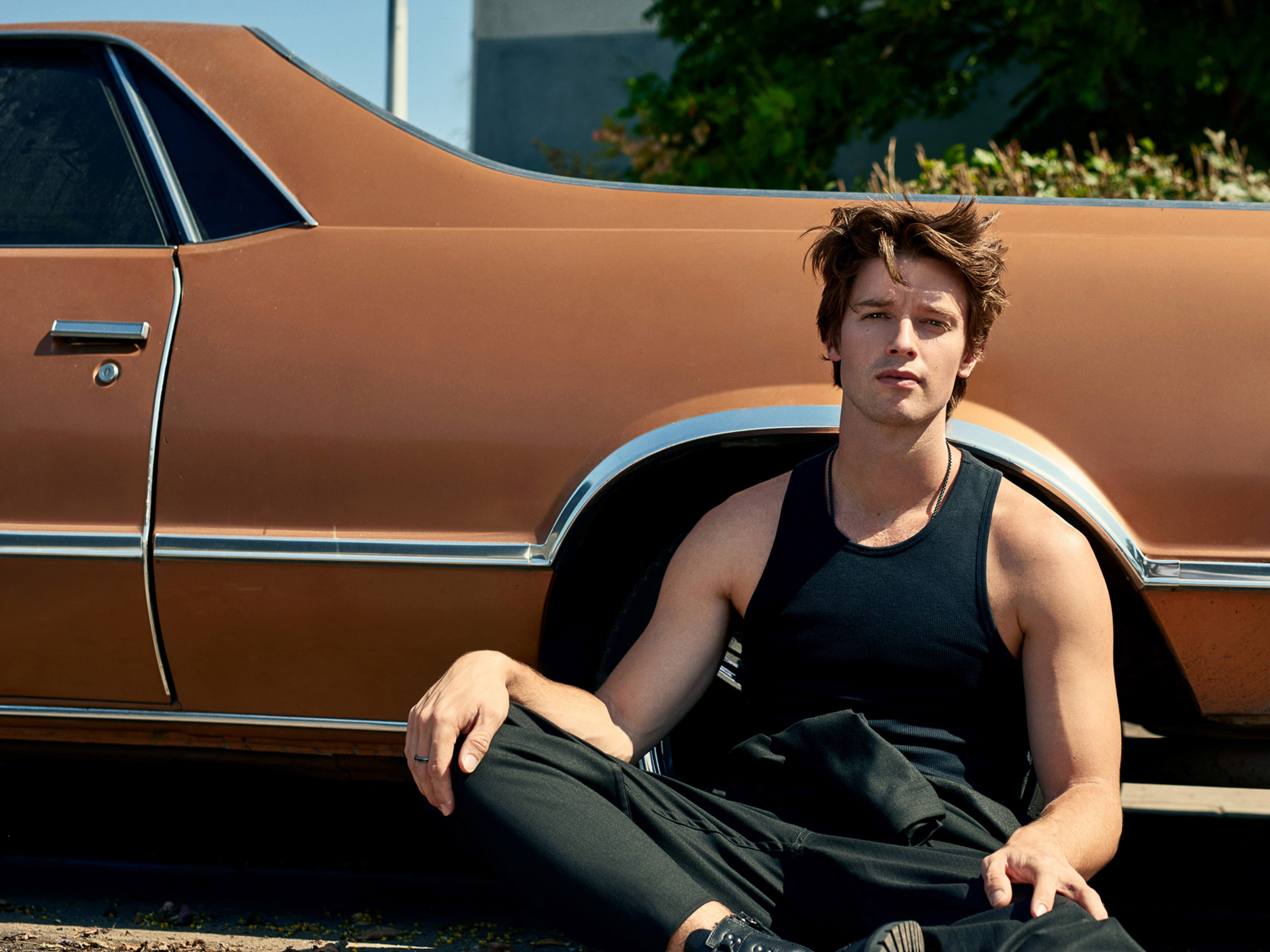 Portrait of a casually dressed young adult male seated by a classic copper car, gazing into the distance with a neutral expression.