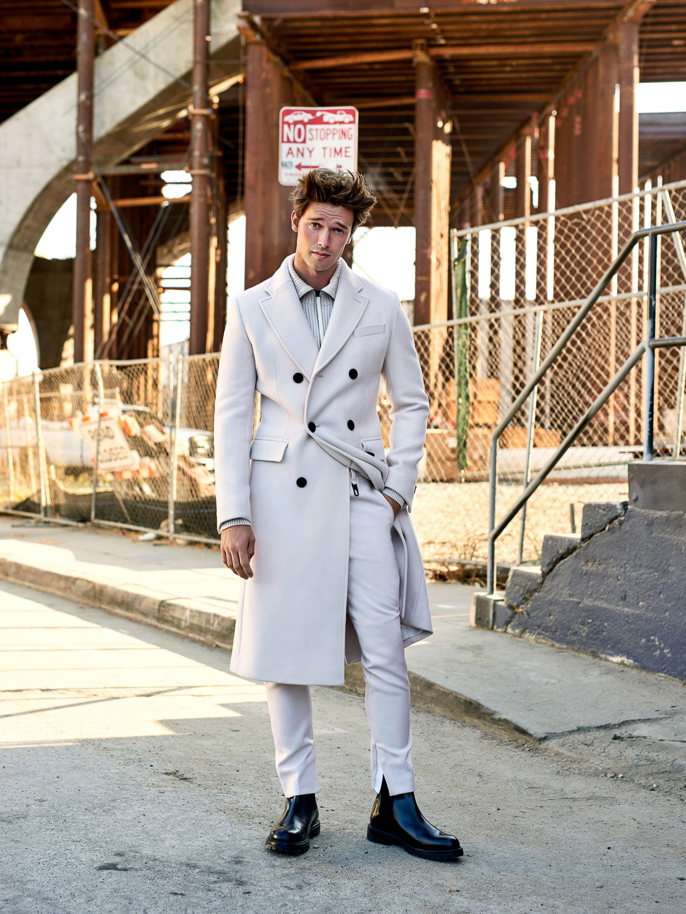 A confident man in a long white coat poses in an urban setting with a concrete road, stairs, and metal beams under a construction site.