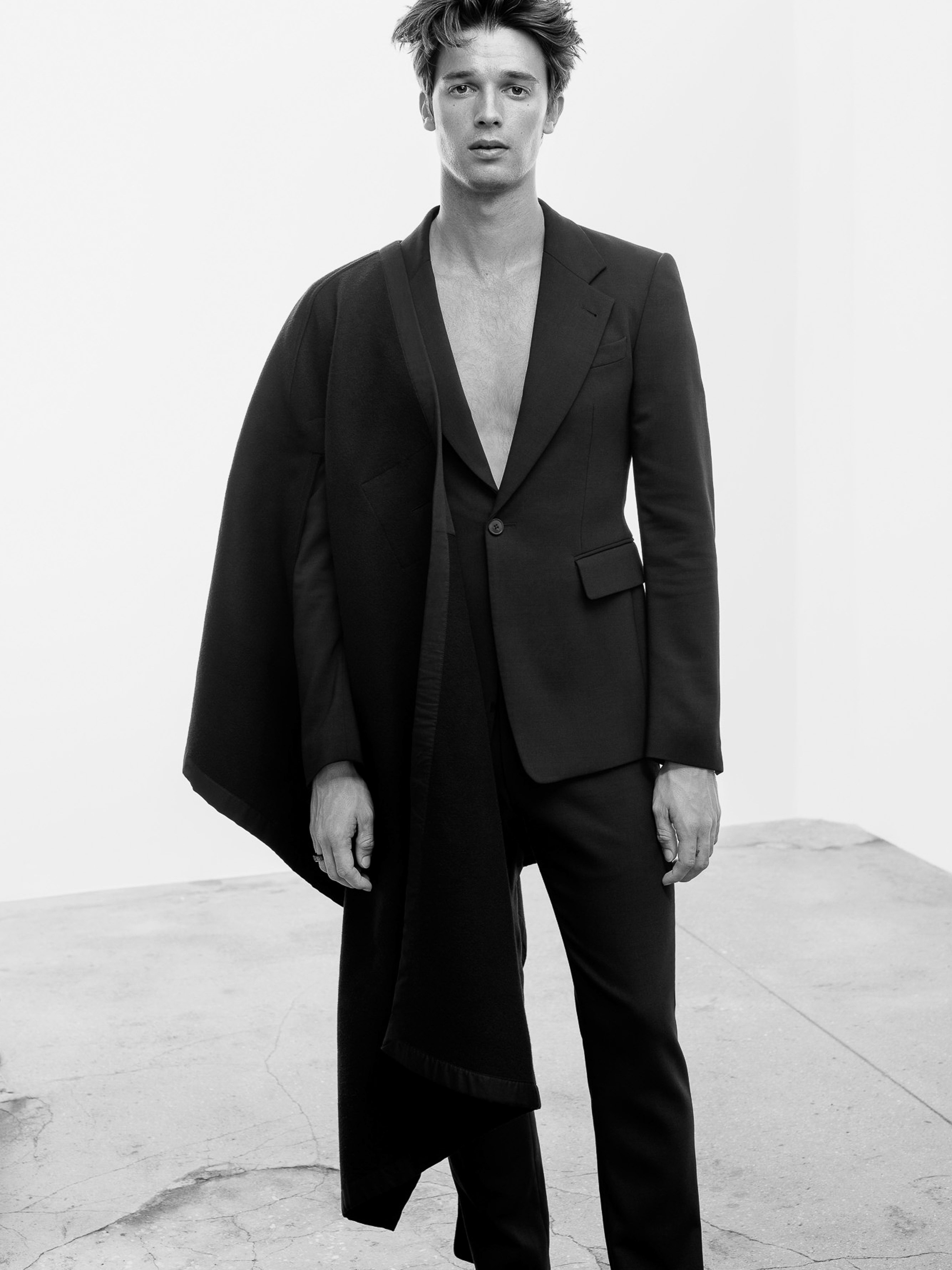 Black-and-white fashion shot of a young man in black suit with open jacket standing in photo studio with concrete floor.