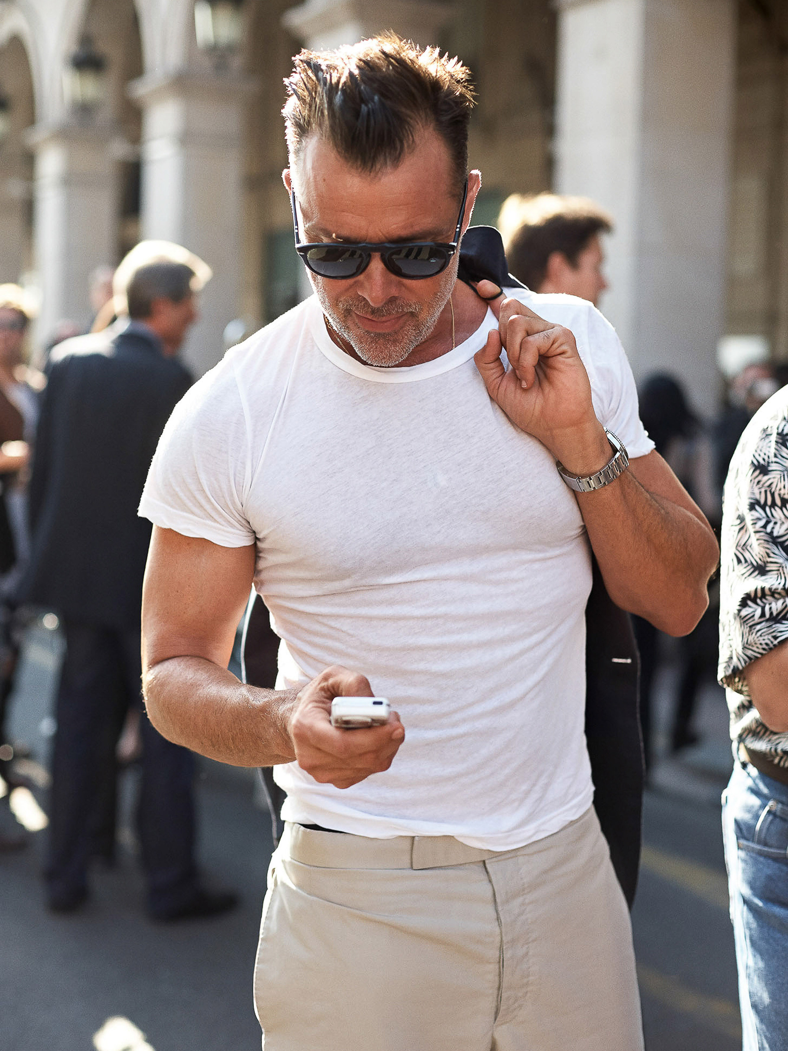 Adult male in a white t-shirt and khaki pants holding a cell phone in a crowd of people outdoors during the day in an urban area.