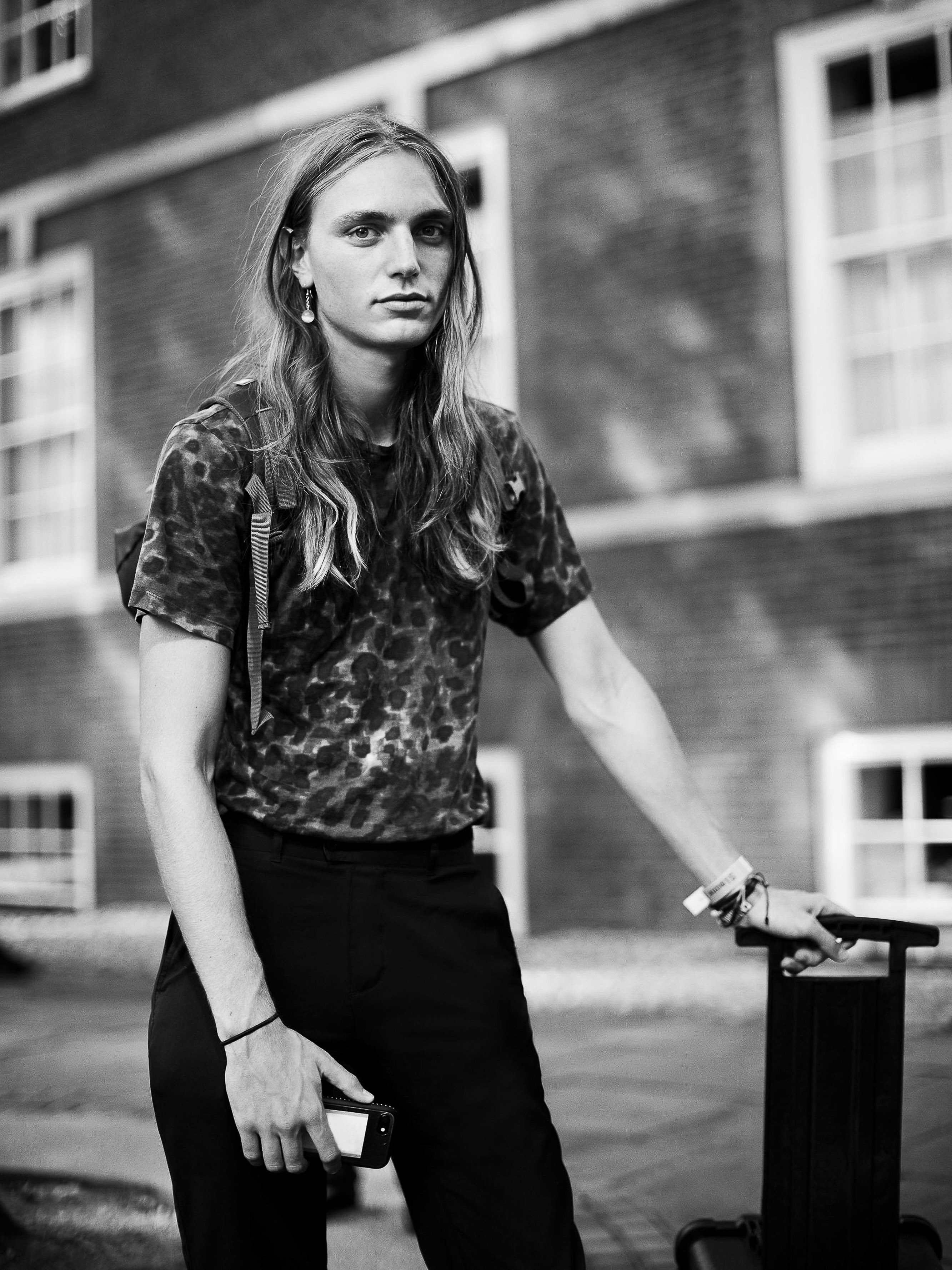 Monochrome portrait of a young adult leaning on a suitcase. The subject has long hair, dark pants, and a tie-dye t-shirt.