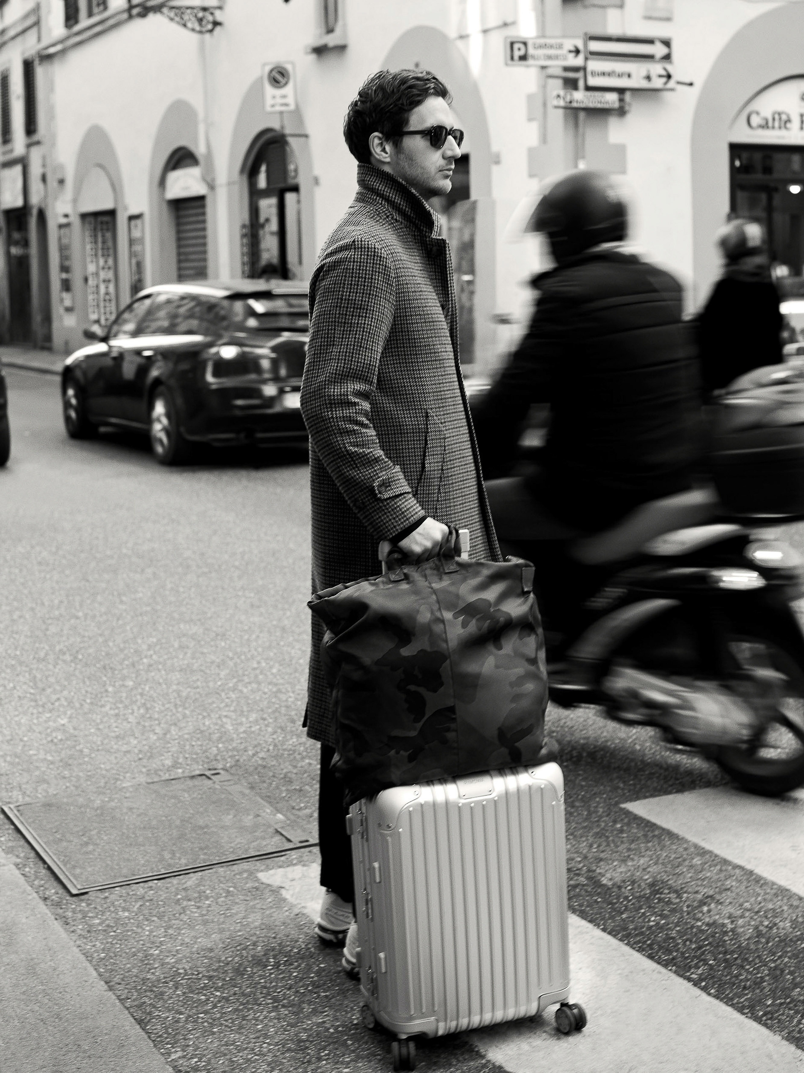 Monochrome portrait of a man in a coat standing with luggage on a city street near a crosswalk, captured at eye-level perspective.