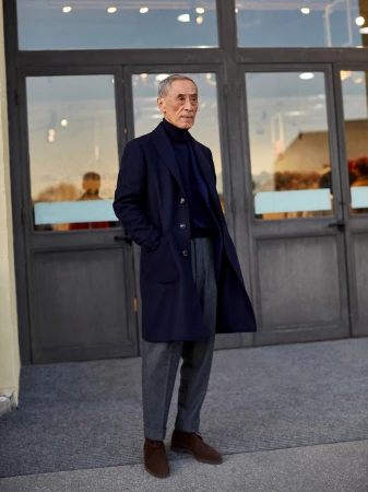 Elegant older man in a navy coat and gray pants stands outside with his hands in his pockets in front of grey paneled doors.