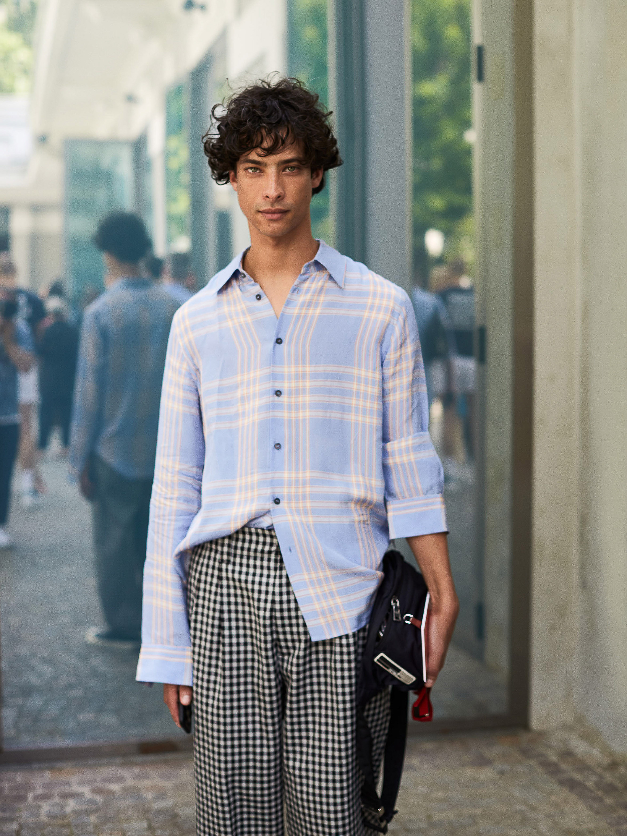 Portrait of a young adult male outdoors, wearing a blue plaid shirt and black and white checkered pants, holding a black shoulder bag.
