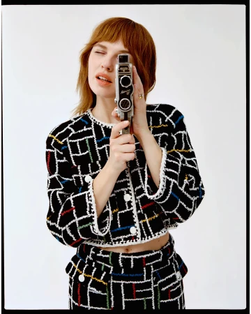 Woman with auburn hair holds a vintage Super 8 film camera, wearing a black and white patterned jacket and skirt against a white backdrop.