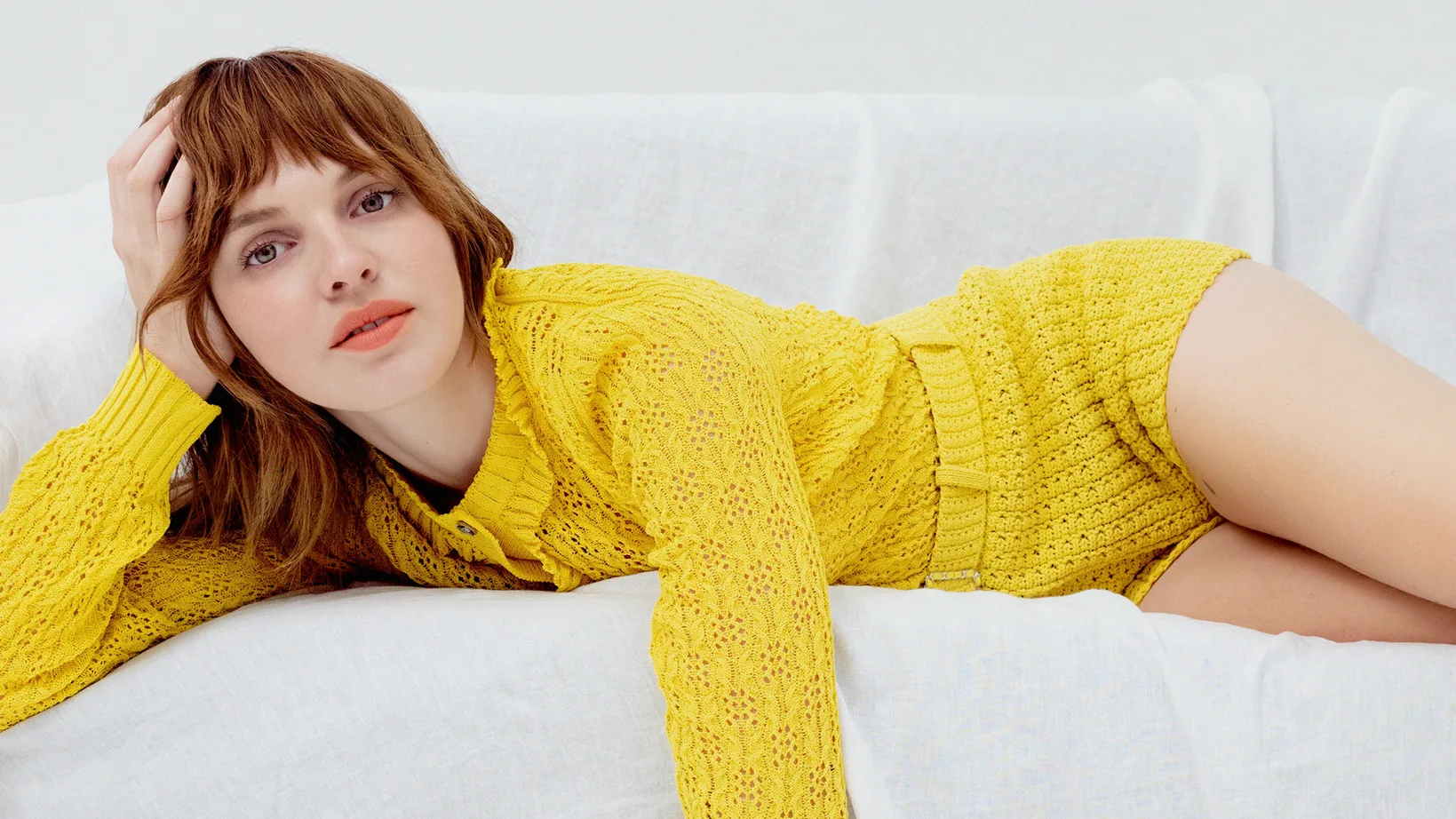A woman with red hair lies on a white surface, wearing a yellow knit outfit and striking a pose, in an indoor setting.