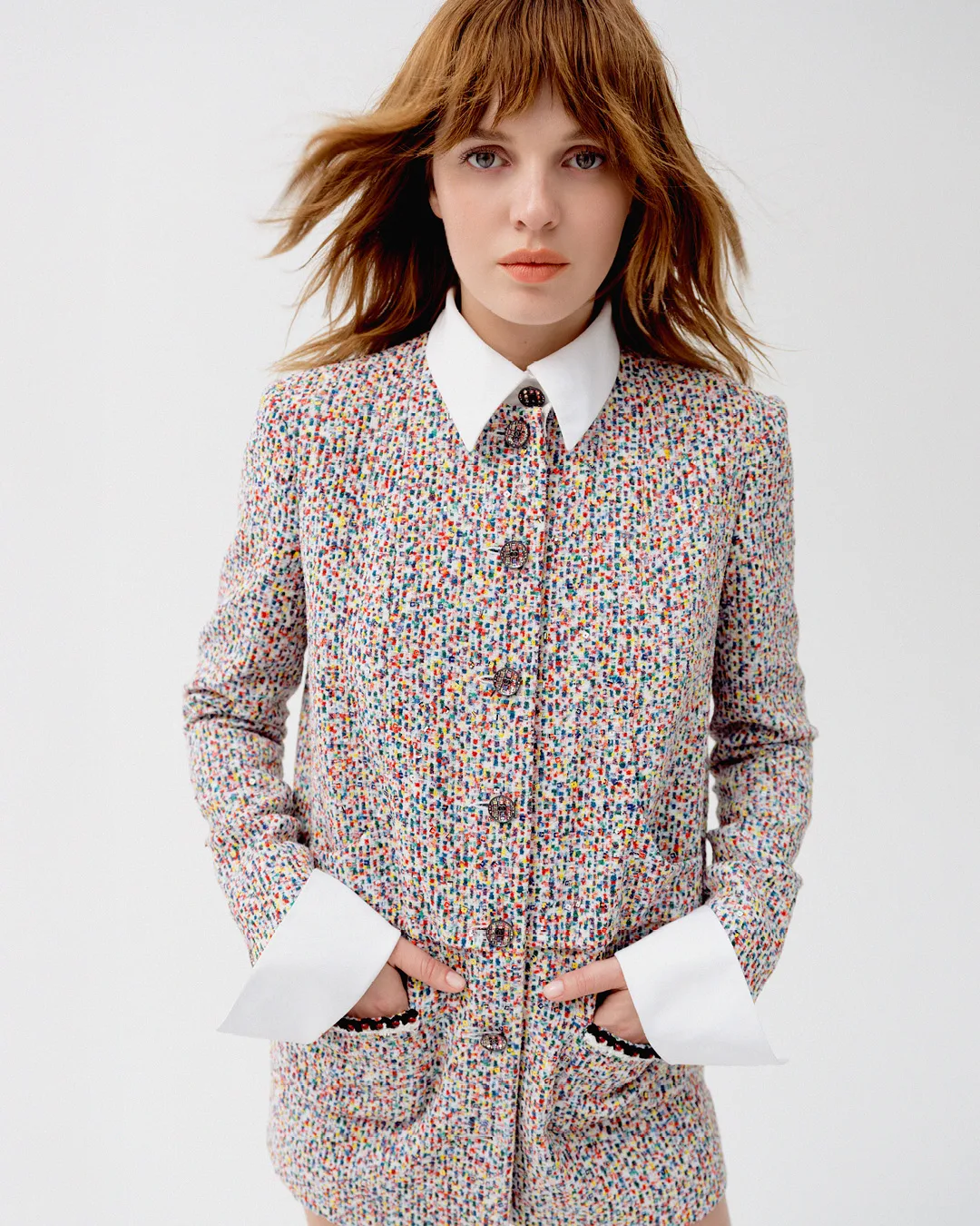 Headshot of a fashionable adult woman with auburn hair wearing a multicolored tweed jacket with white collar in a studio setting.