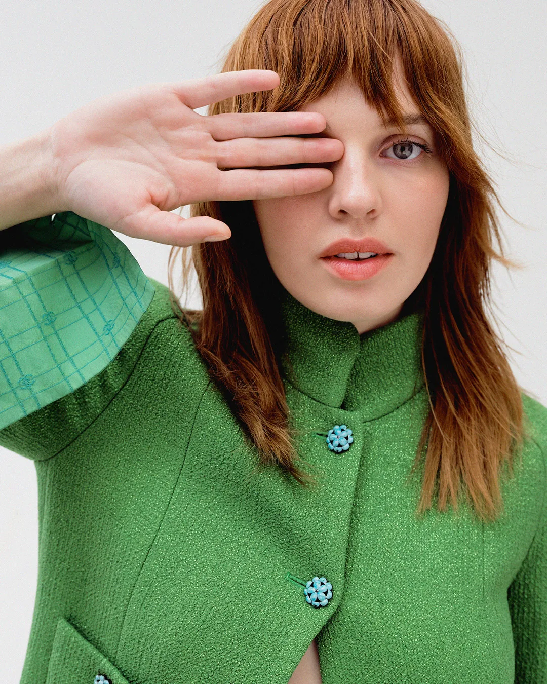 A young woman with red hair poses in a green jacket, partially covering her face with her hand, in a studio shot on a white background.