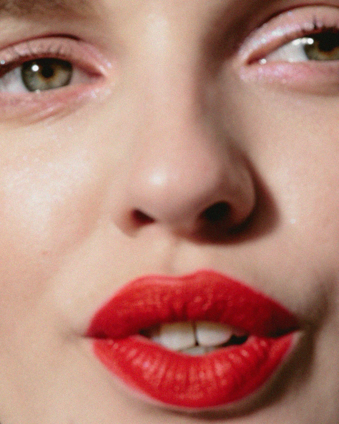 Close-up of a woman's face with bright red lipstick, fair skin, and green eyes, capturing makeup details and facial features in soft lighting.