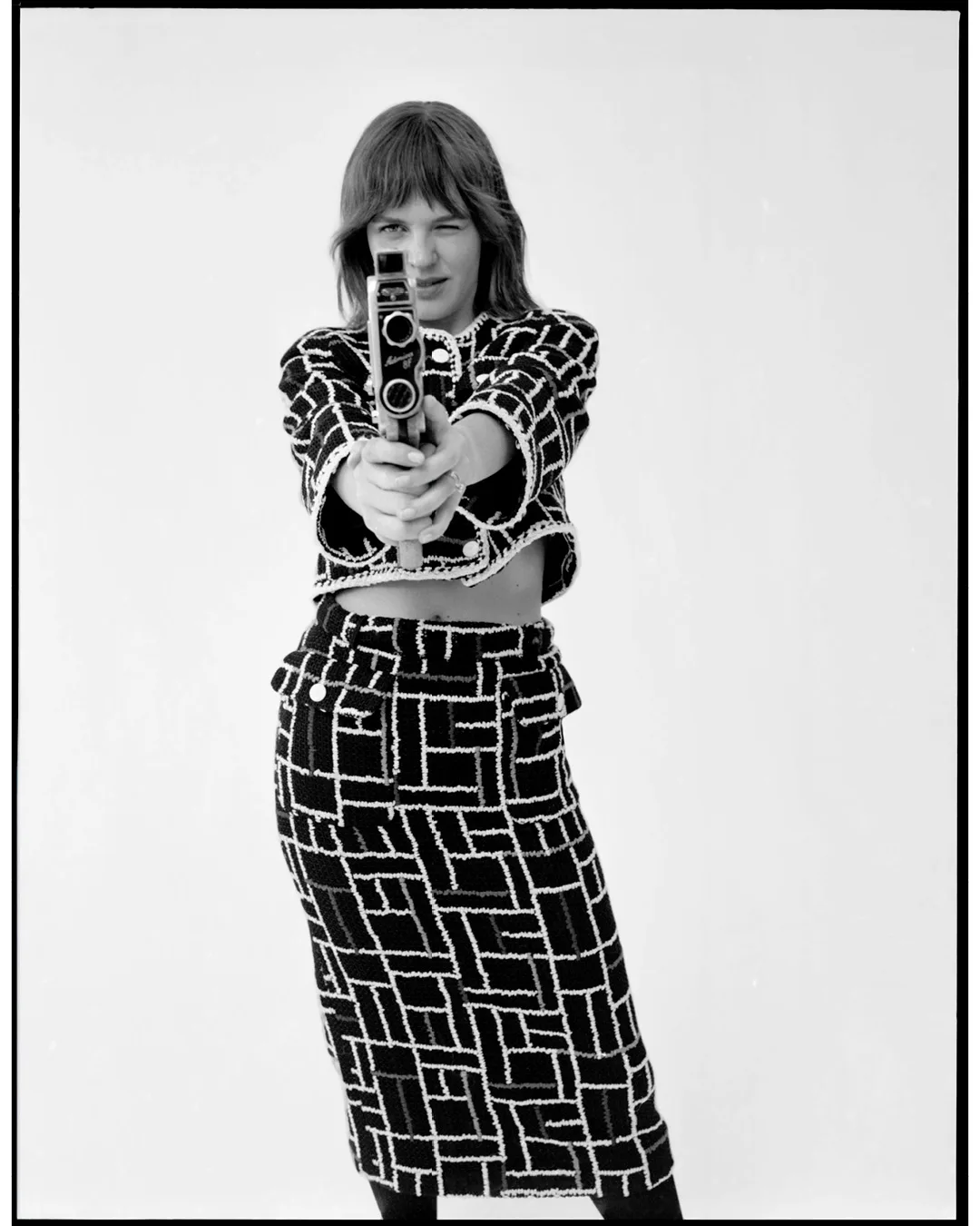 Woman poses in a black and white patterned suit, pointing an old Super 8 camera directly at the viewer, indoors against a white background.