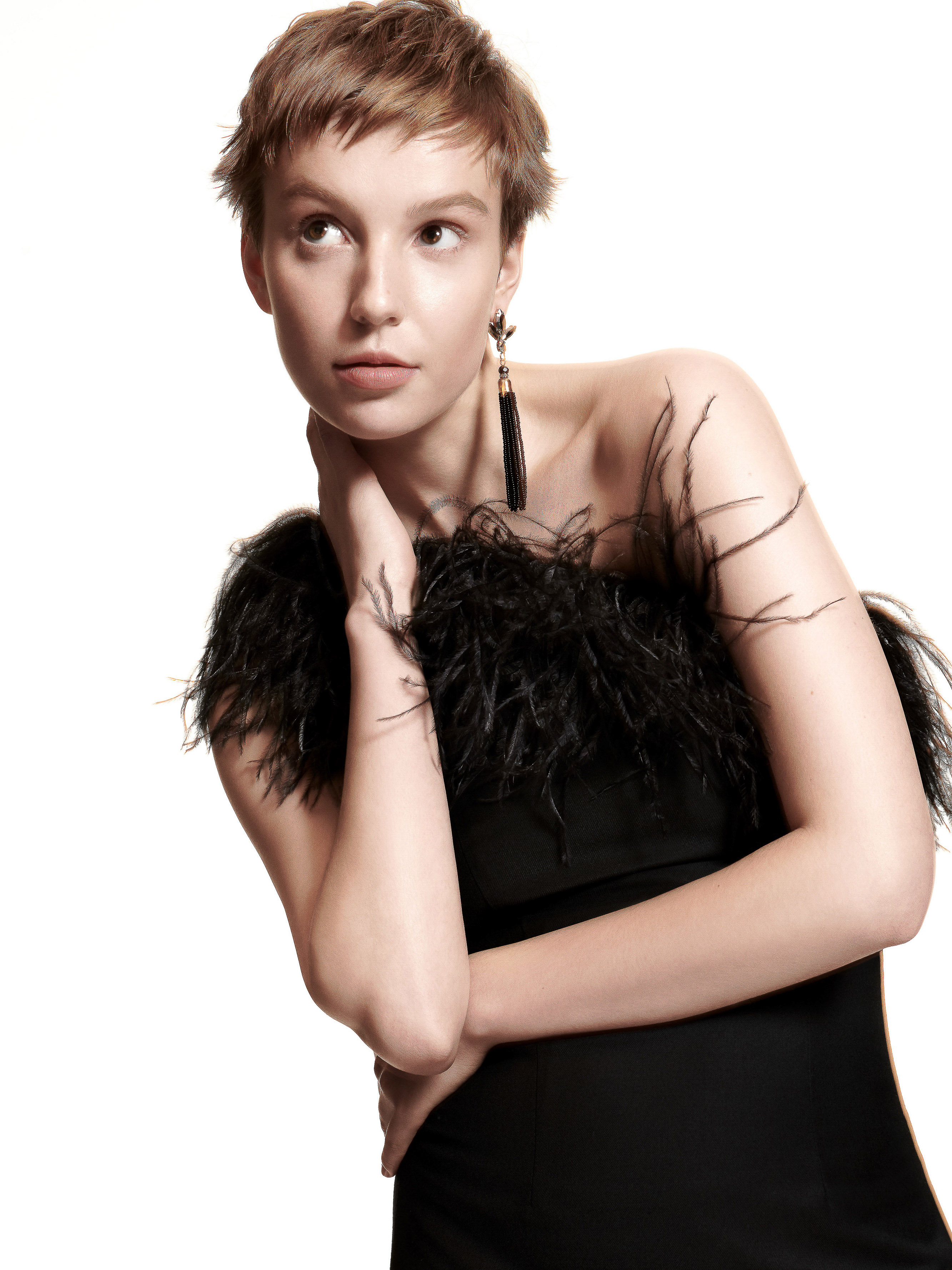 Portrait of a woman in a black dress with feather trim. She's wearing statement earrings and poses in front of a white background.