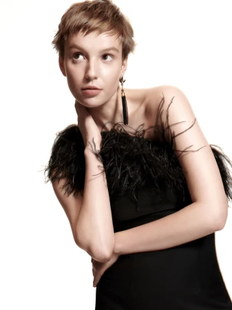 Portrait of a woman in a black dress with feather trim. She's wearing statement earrings and poses in front of a white background.