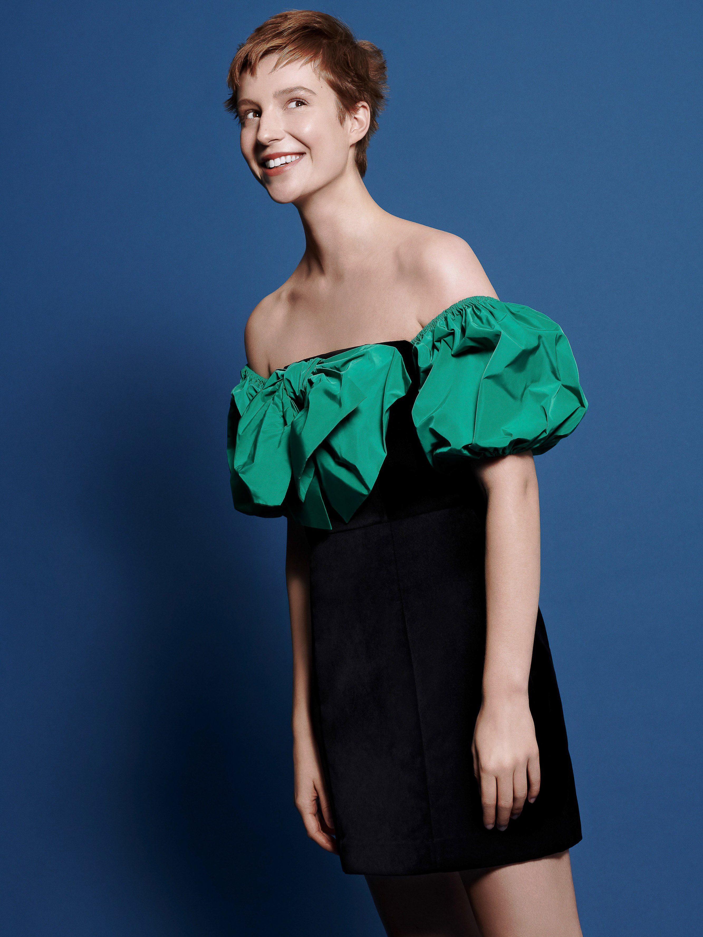 Smiling woman in an off-the-shoulder green and black dress against a solid blue background, looking up and to the left.