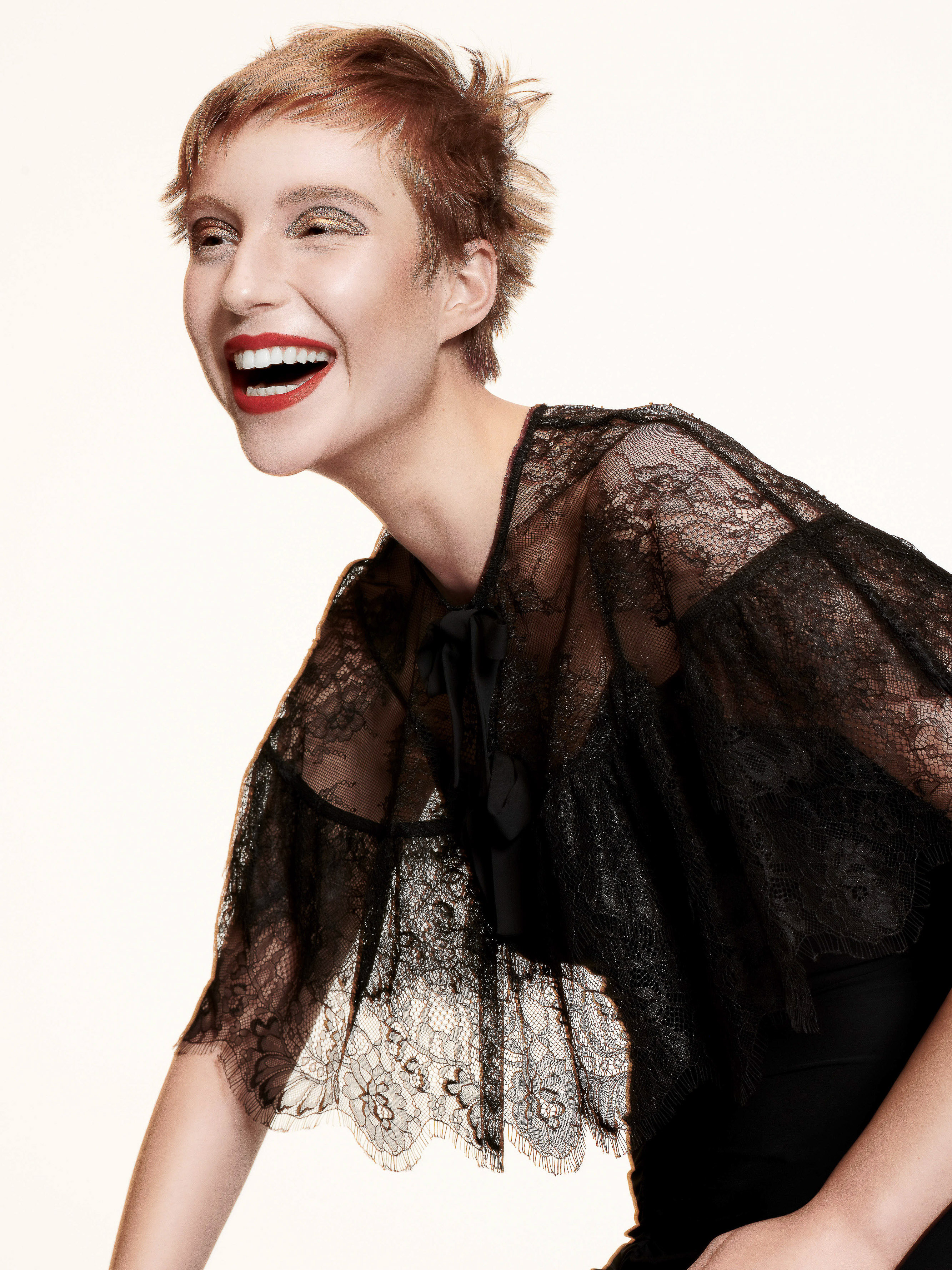 Woman with short red hair laughing joyfully while wearing a black sheer lace cape against a white studio background.