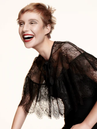 Woman with short red hair laughing joyfully while wearing a black sheer lace cape against a white studio background.