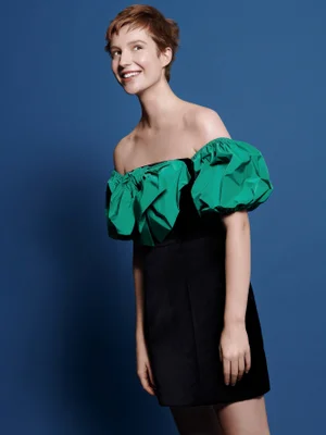 Smiling woman with short brown hair wearing an off-the-shoulder black dress with green puffed sleeves against a blue backdrop.
