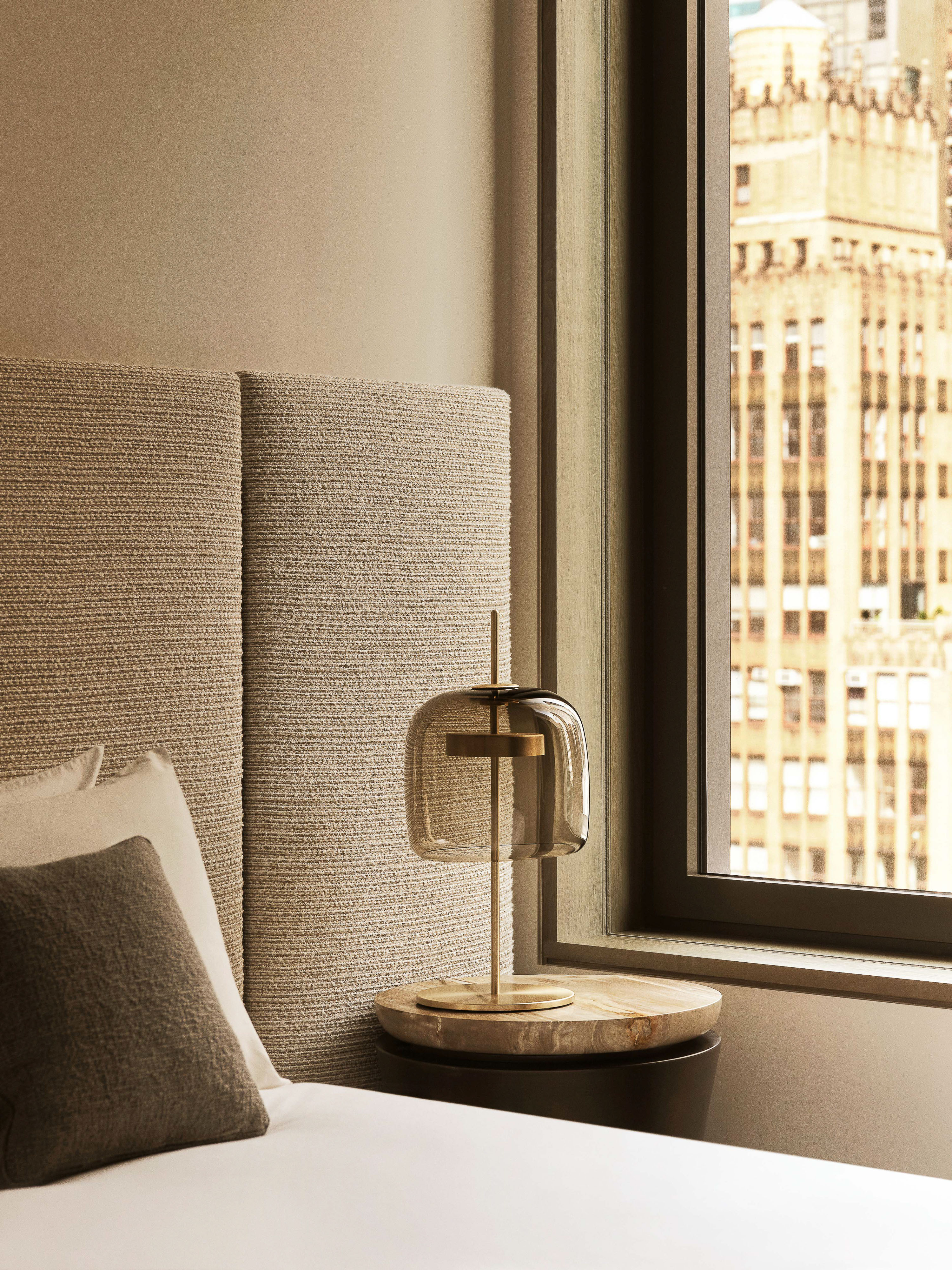 Modern bedroom interior showcasing a table lamp on a bedside table next to a textured headboard, with a city view through a window.
