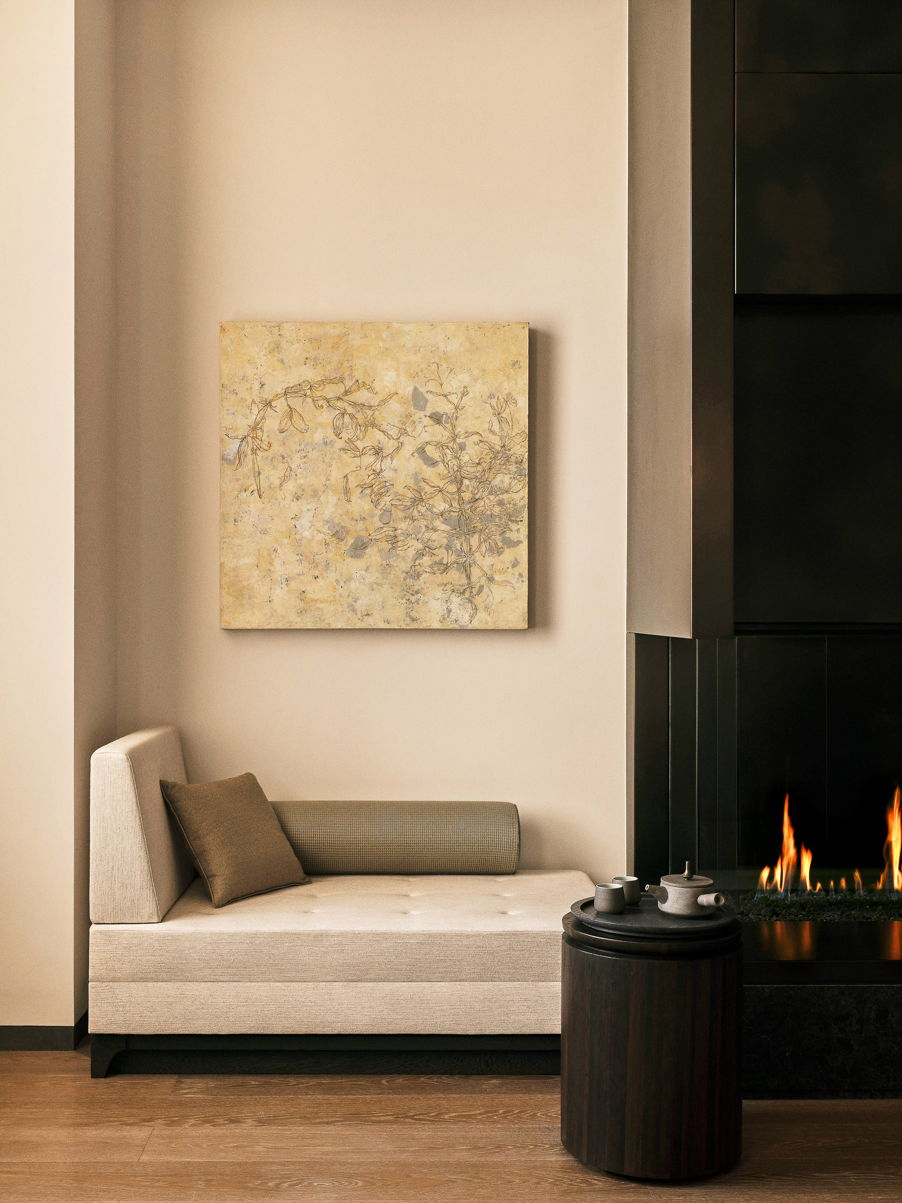 Beige modern interior with daybed, abstract art, black fireplace, and a small round table holding tea set next to wooden floor.