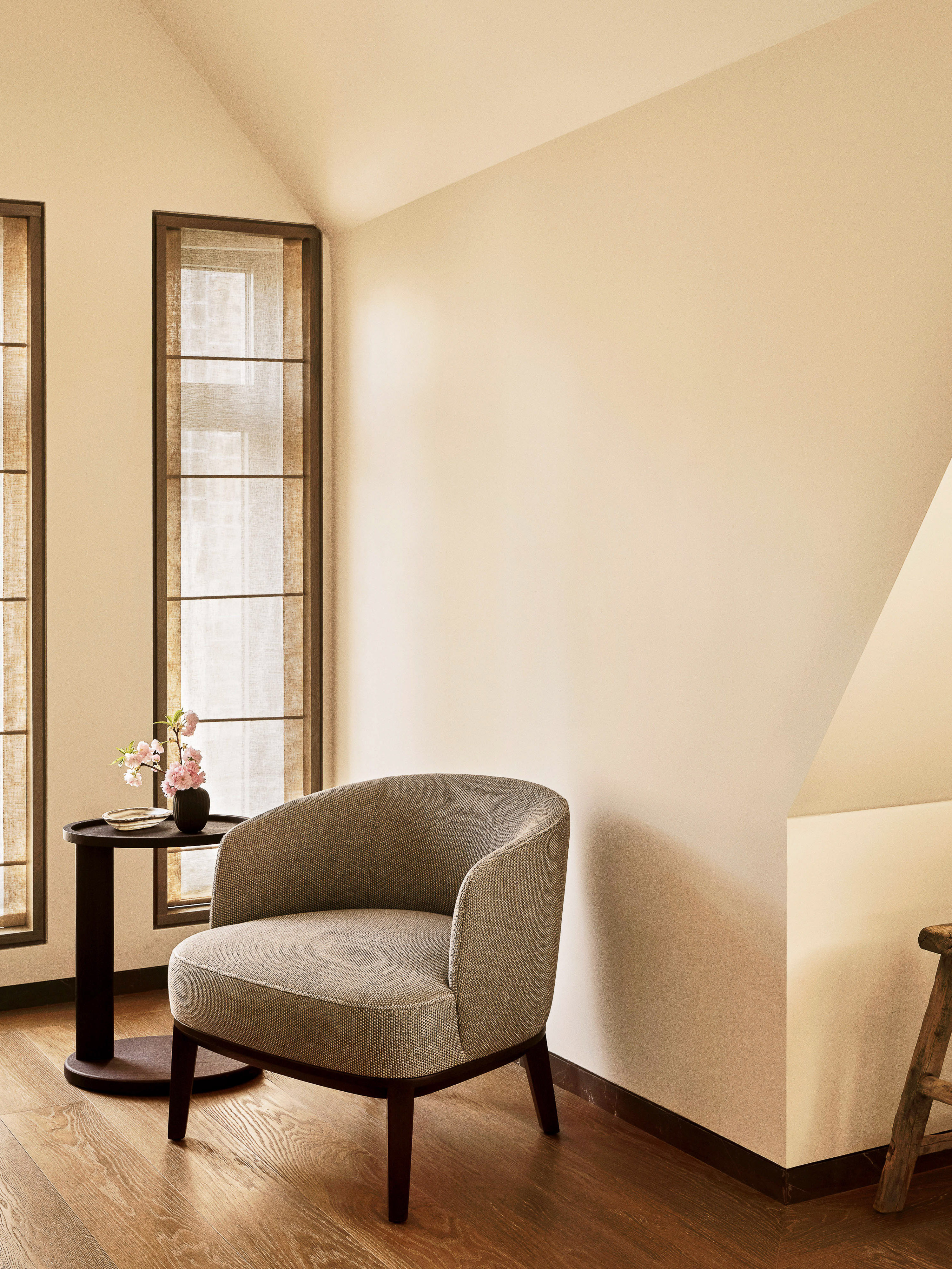 Interior featuring a gray upholstered armchair on a wooden floor, with a side table, flower arrangement, two tall windows, and neutral walls.