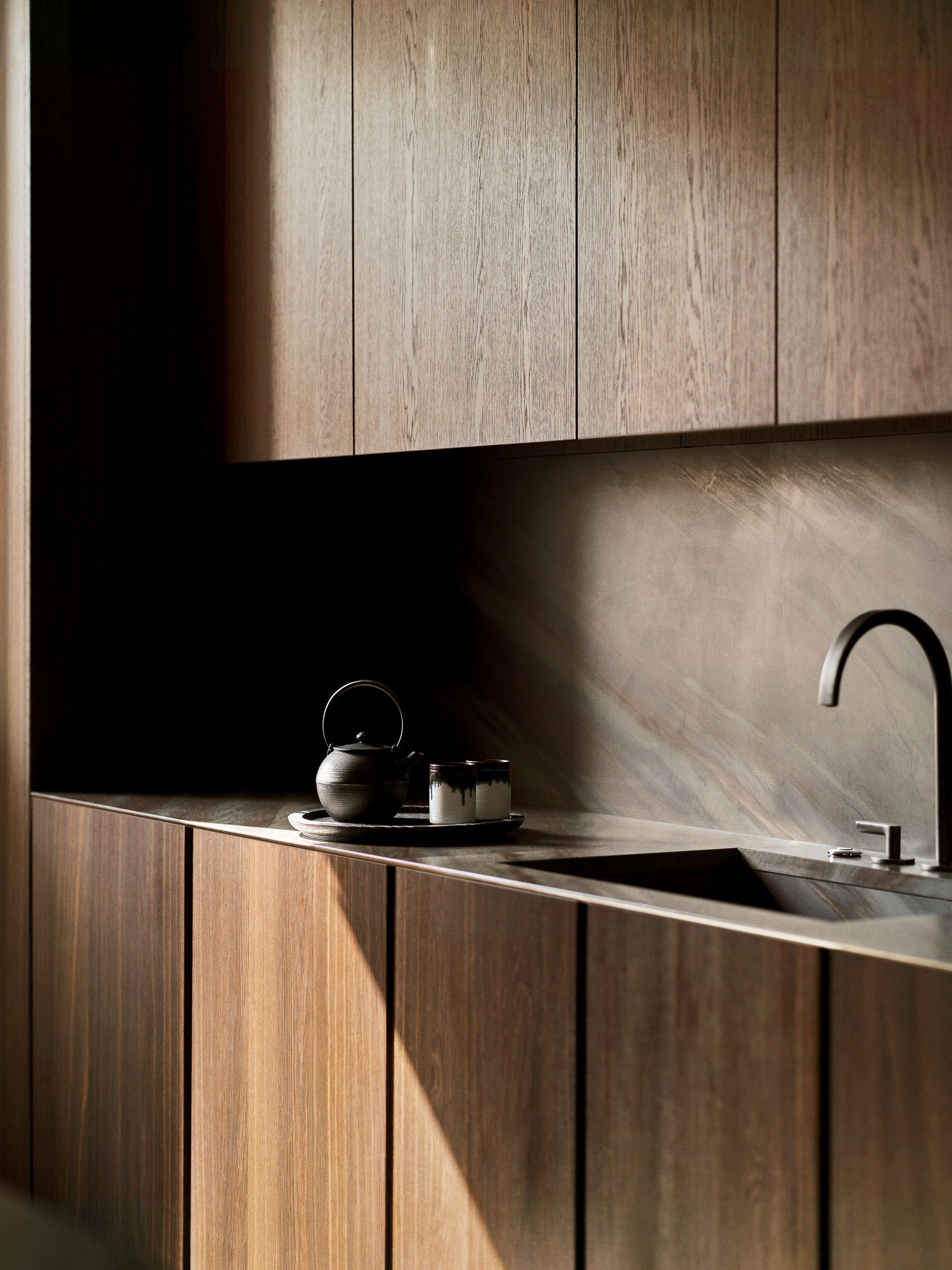 Minimalist kitchen interior featuring dark wood cabinets, a black teapot, and a modern black faucet. Natural sunlight highlights the textures.
