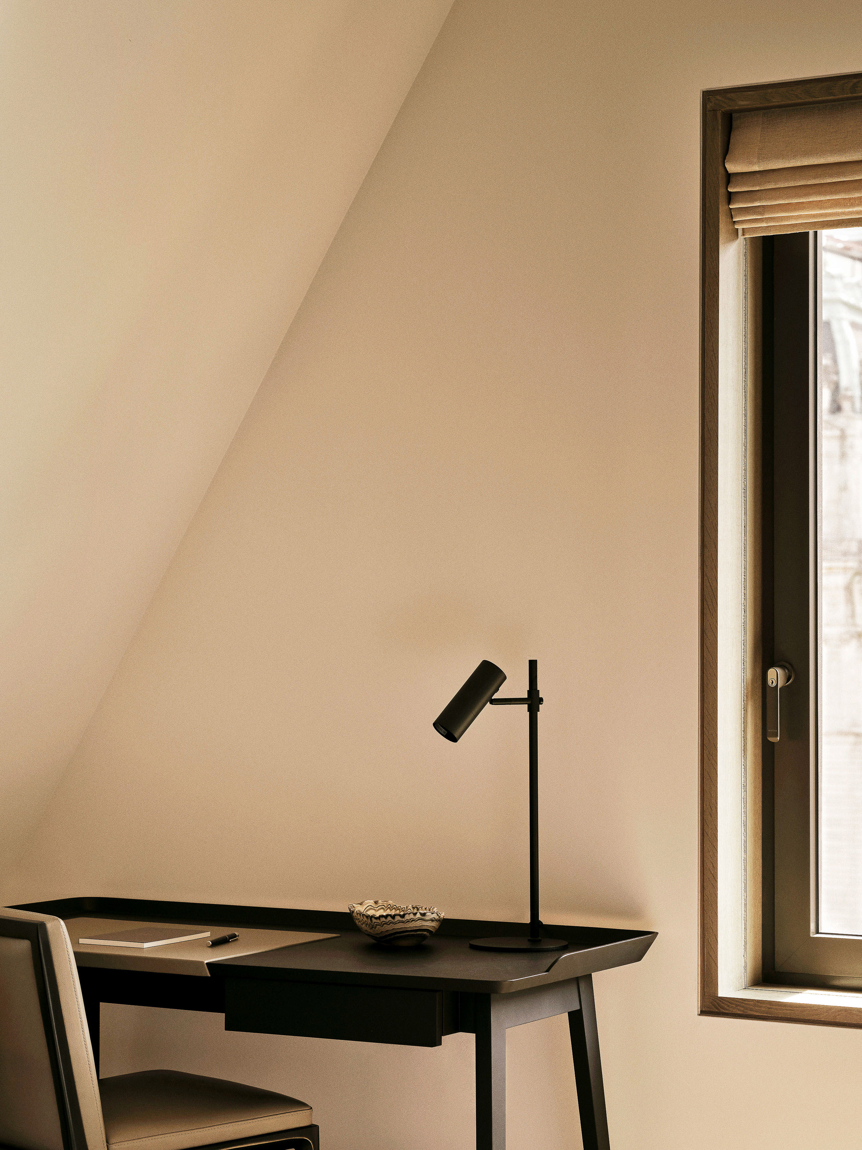 Minimalist interior view of a writing desk with a black lamp, chair, window with blinds, and neutral-toned wall in natural light.