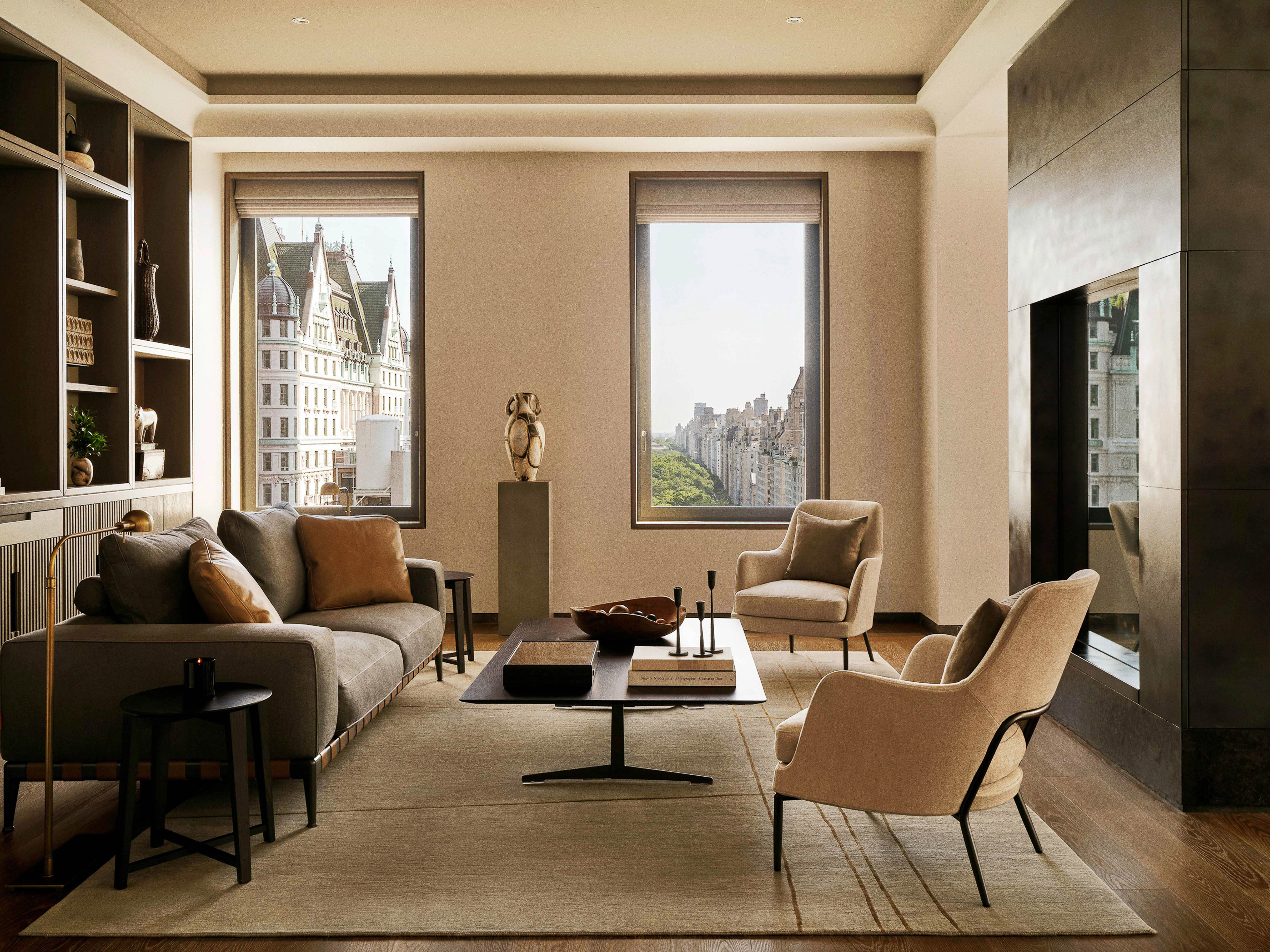 Living room interior featuring a grey sofa with pillows, a dark coffee table, and cream-colored armchairs, bathed in natural light.