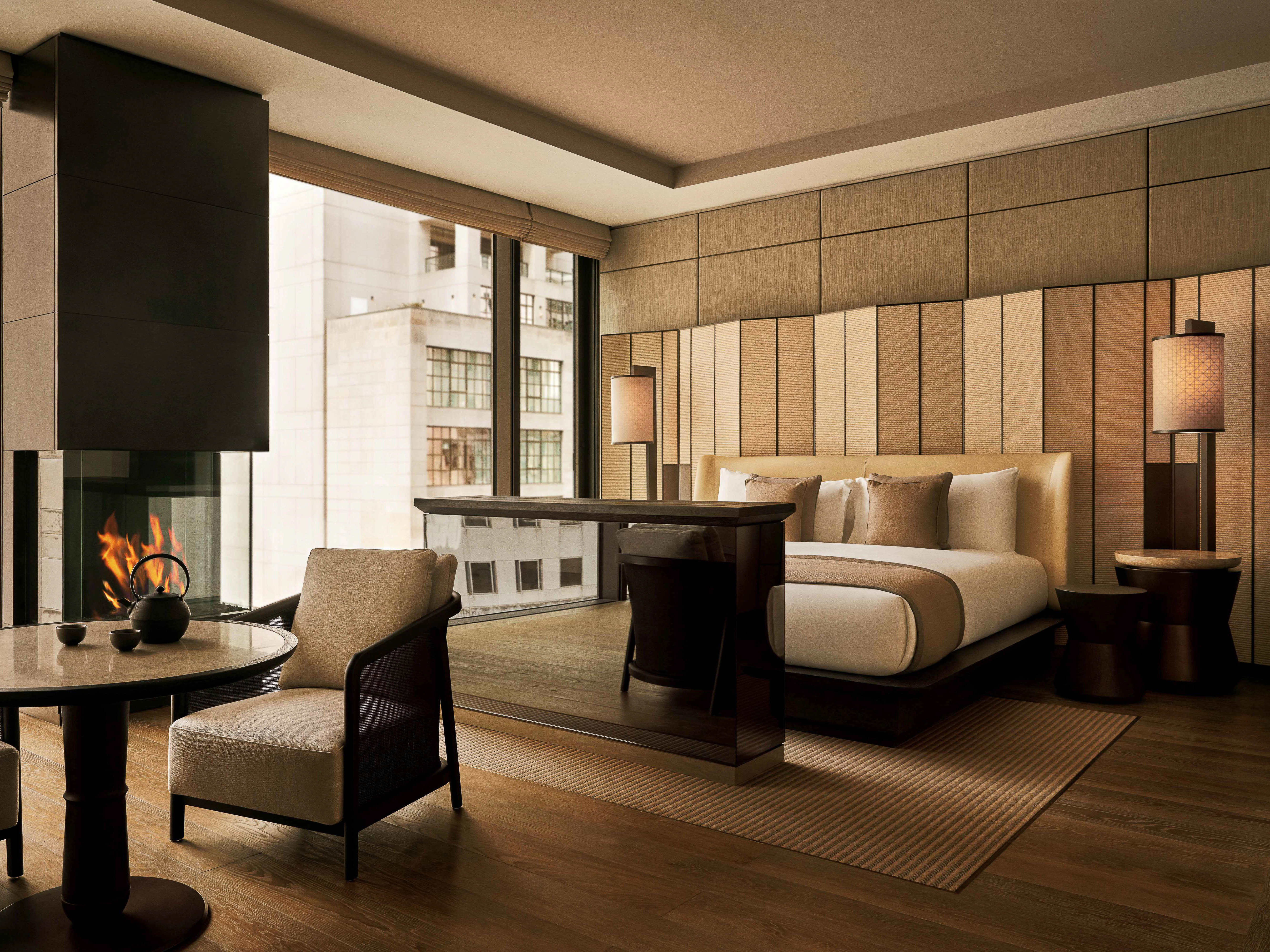 A luxurious hotel room interior features a comfortable bed, fireplace, table with seating, and a large window with a city view.