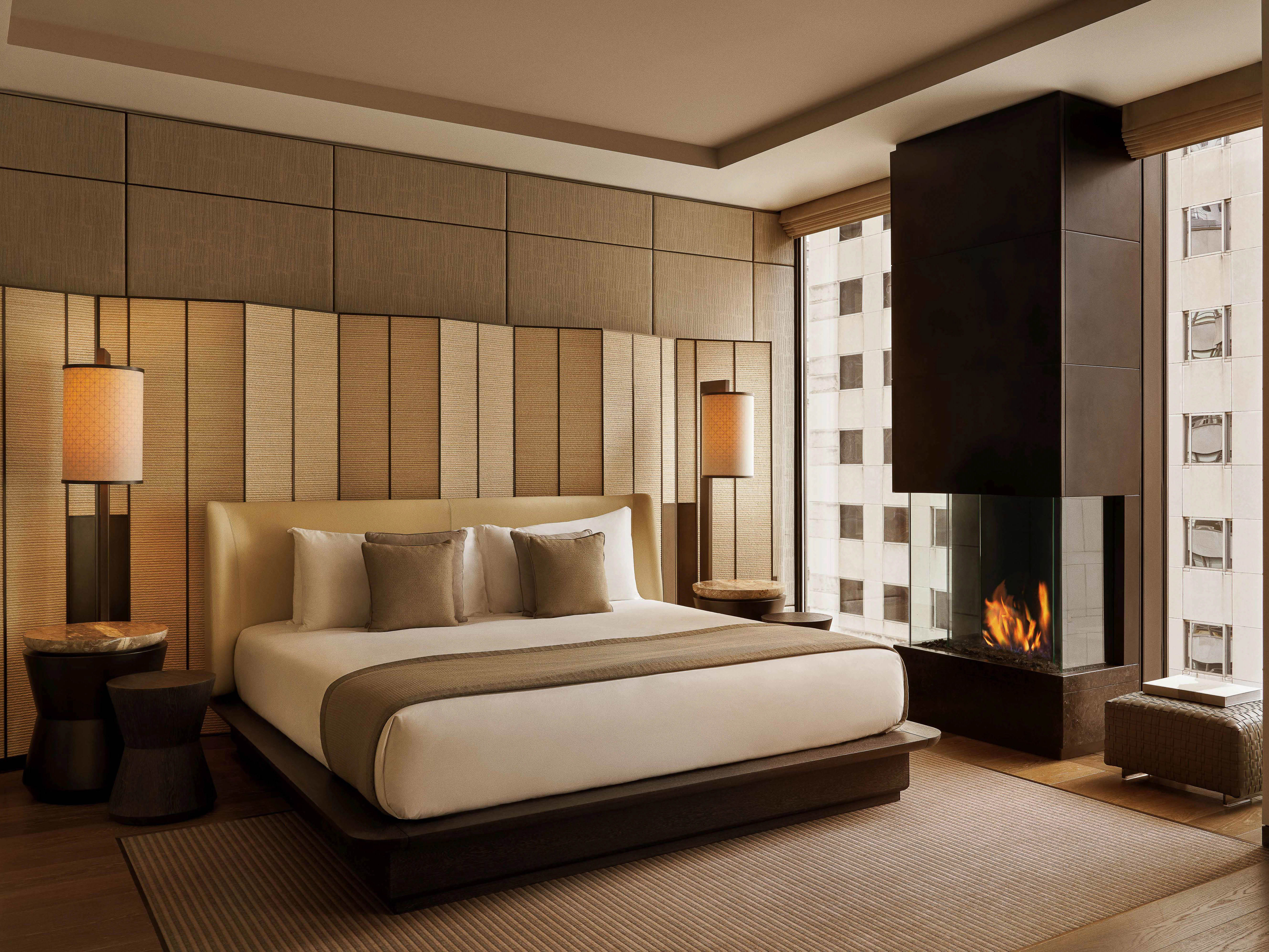 Hotel bedroom interior featuring a large bed with pillows, lamps on side tables, a modern fireplace, and a window with a city view.