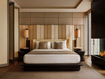 Symmetrical luxury hotel bedroom with a platform bed, white bedding, woven wood headboard, bedside lamps, and a modern fireplace.