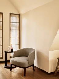 A grey upholstered barrel armchair sits on hardwood flooring beside a small dark round table with pink blossoms in a bright interior.