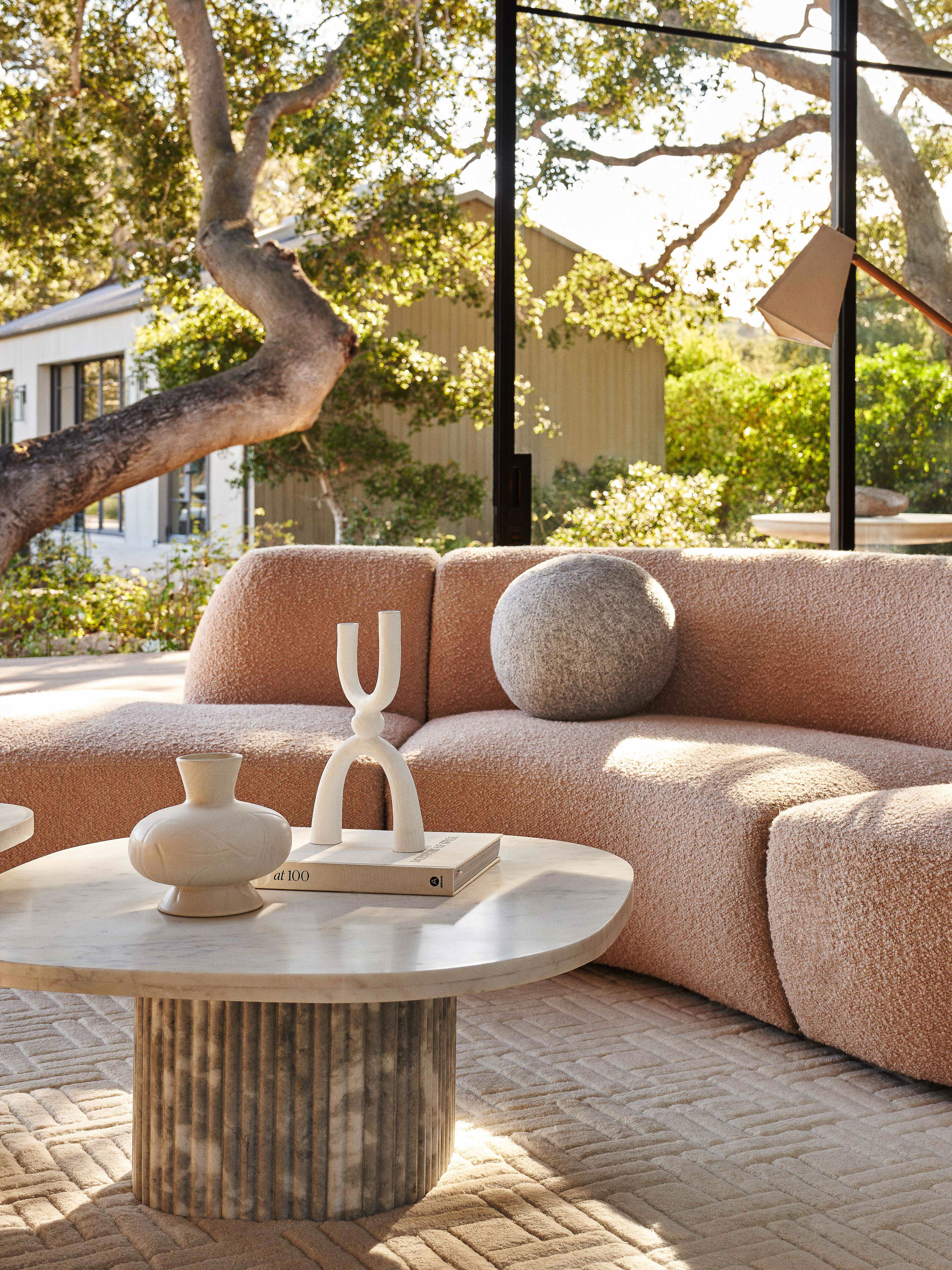 Living room interior featuring a peach-colored sectional sofa, round marble coffee table, decor objects, and large windows overlooking greenery.