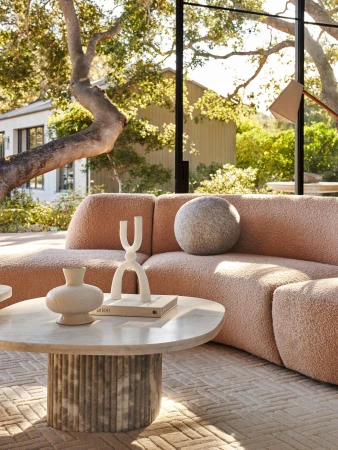 Living room interior featuring a peach-colored sectional sofa, round marble coffee table, decor objects, and large windows overlooking greenery.