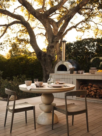 Outdoor dining scene with a round table set for a meal, surrounded by wooden chairs and a pizza oven in a lush backyard setting.