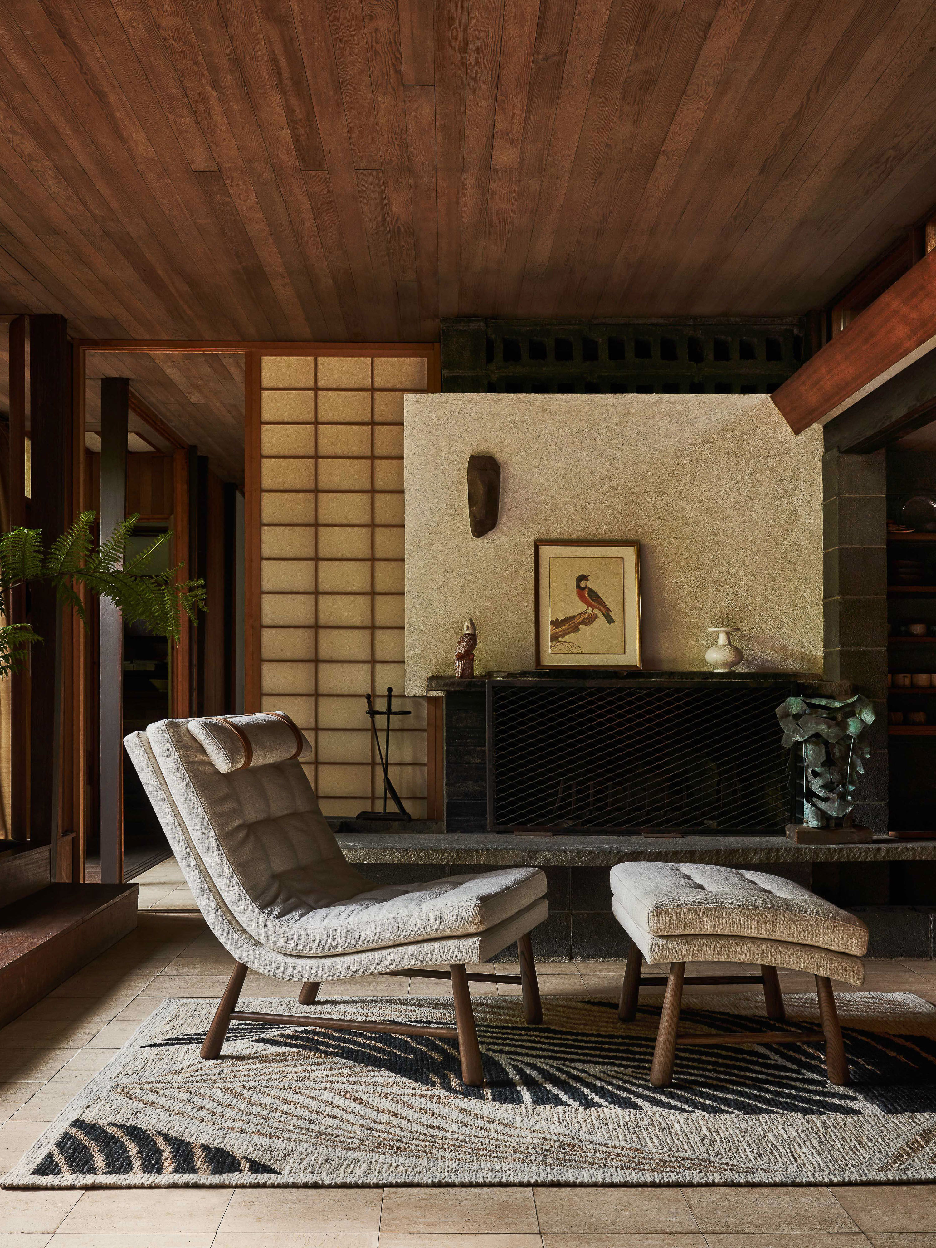 Interior view of a neutral-toned modern living room with a lounge chair, footstool, graphic rug, and decorative fireplace.